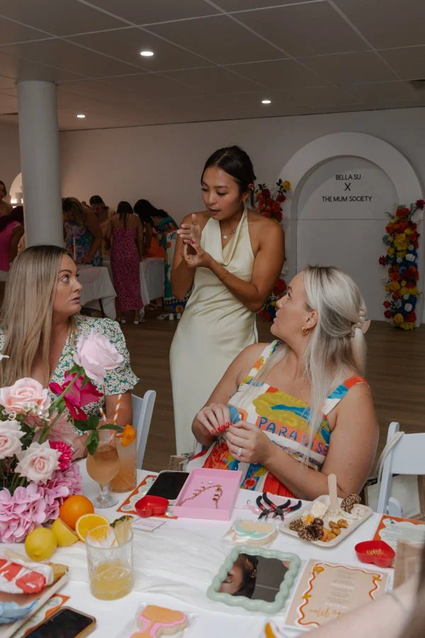 Three women at a celebration table with flowers, drinks, and desserts, with a decorated background and people in the background.