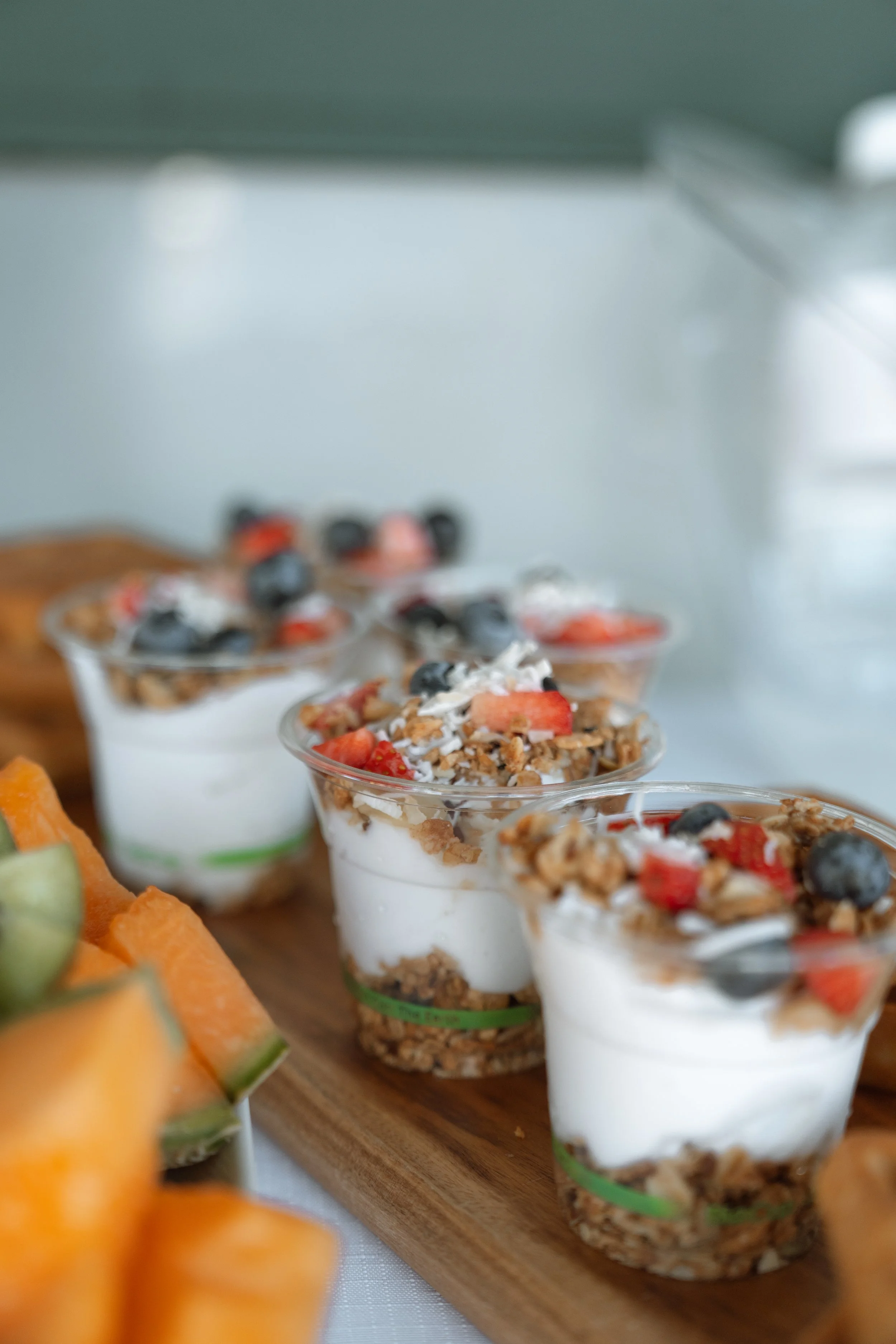 Several parfait desserts layered with granola, yogurt, and fresh berries on a wooden tray, with sliced melon in the foreground.