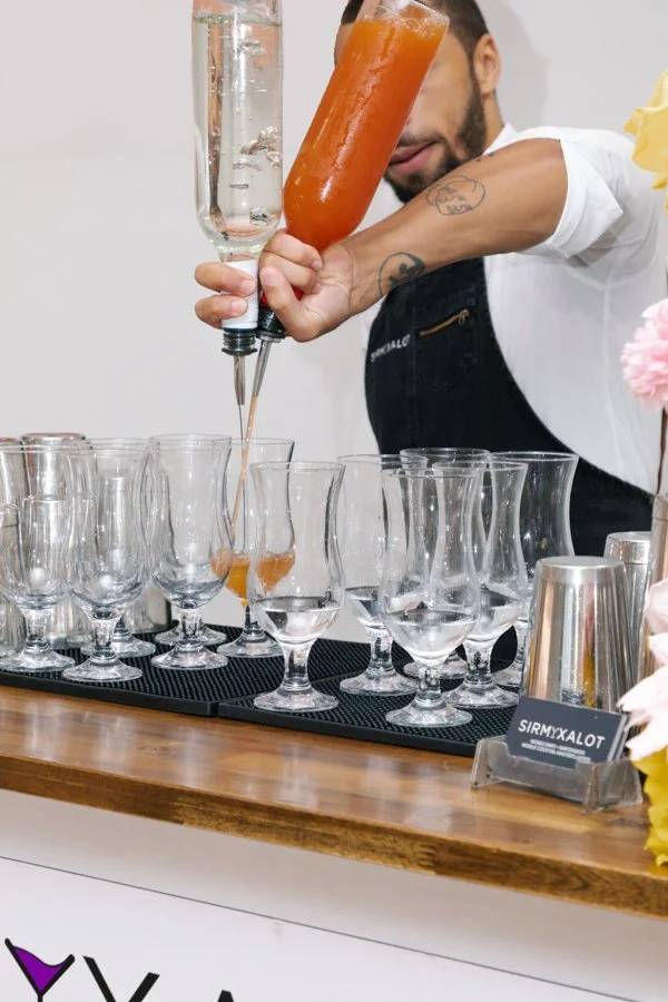 Bartender pouring a layered drink into glassware, with many empty glasses lined up on a bar counter.