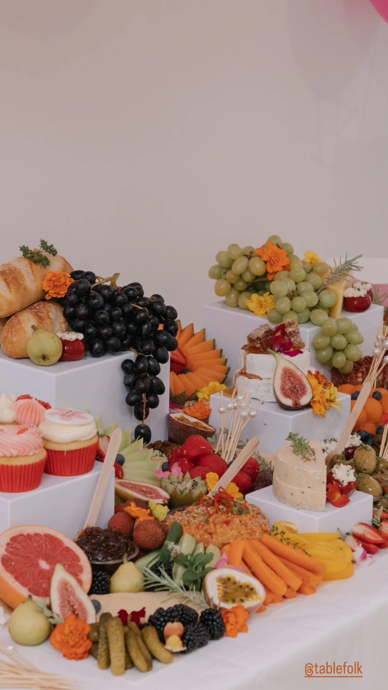 A colorful display of assorted fruits and snacks, including grapes, figs, strawberries, oranges, and honey, along with cupcakes, cheeses, and decorative flowers on white platforms, arranged on a white table.