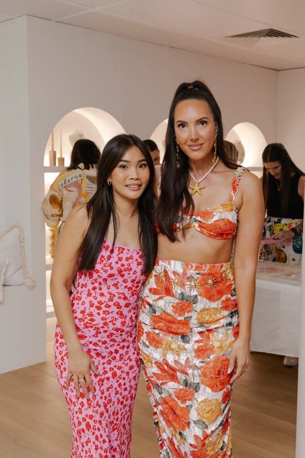Two women standing side by side in a bright, modern room with white walls and arched niches, dressed in colorful floral dresses, smiling at the camera.