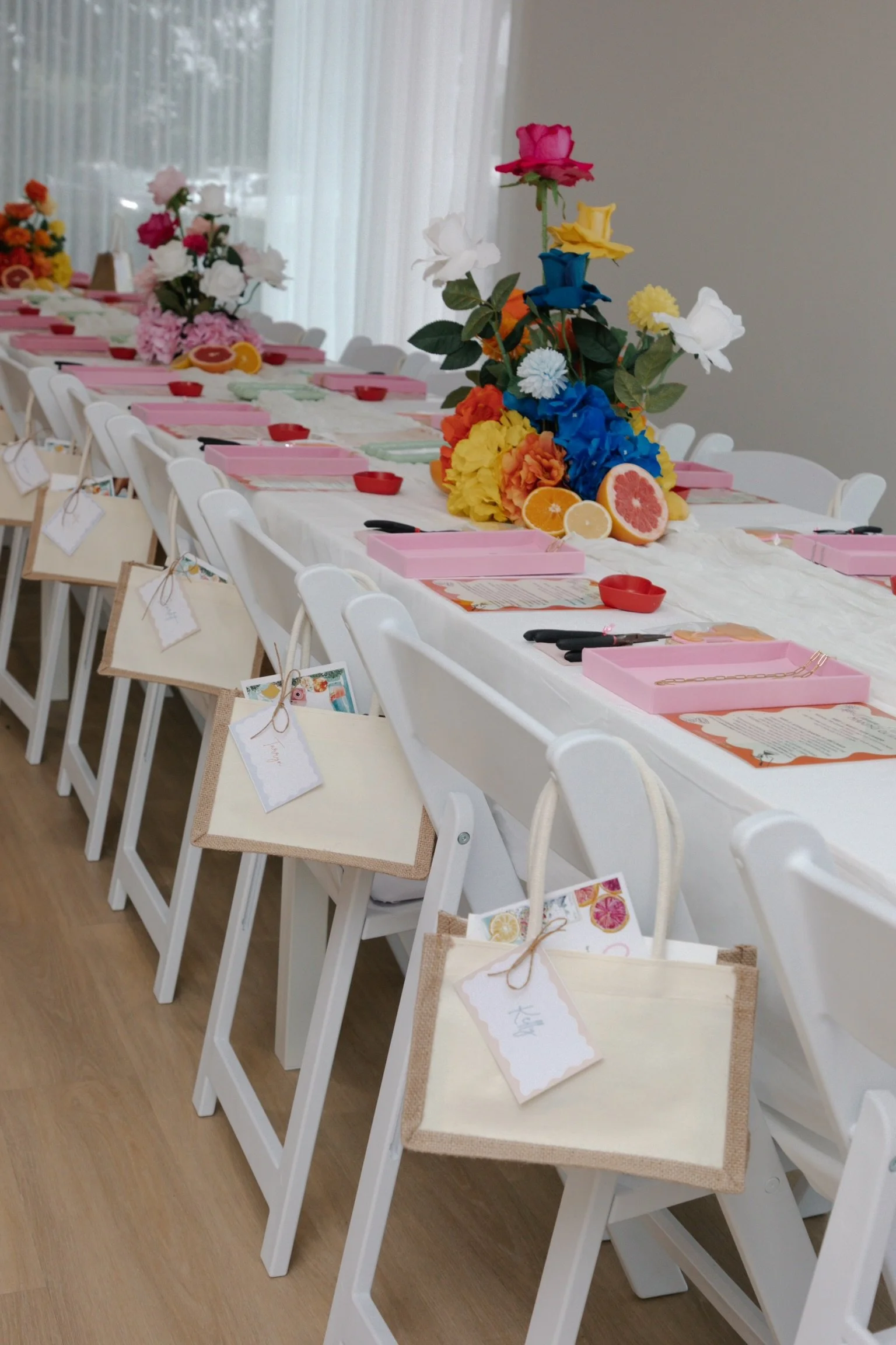 A long table decorated with vibrant floral arrangements, pink boxes, and place settings for a party or celebration, with chairs tagged with name cards hanging on the back.