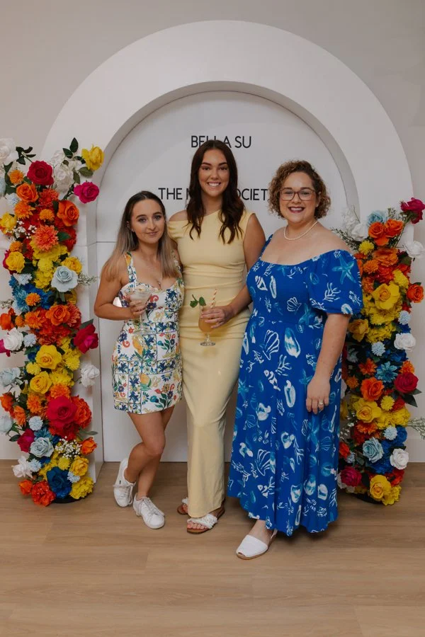 Three women standing in front of a floral arch at an event, smiling and holding drinks.