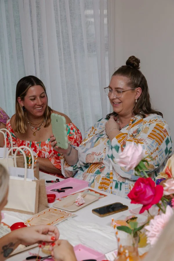 Two women sitting at a table, smiling, one holding a phone and showing an image to the other, with decorated gifts and flowers on the table.
