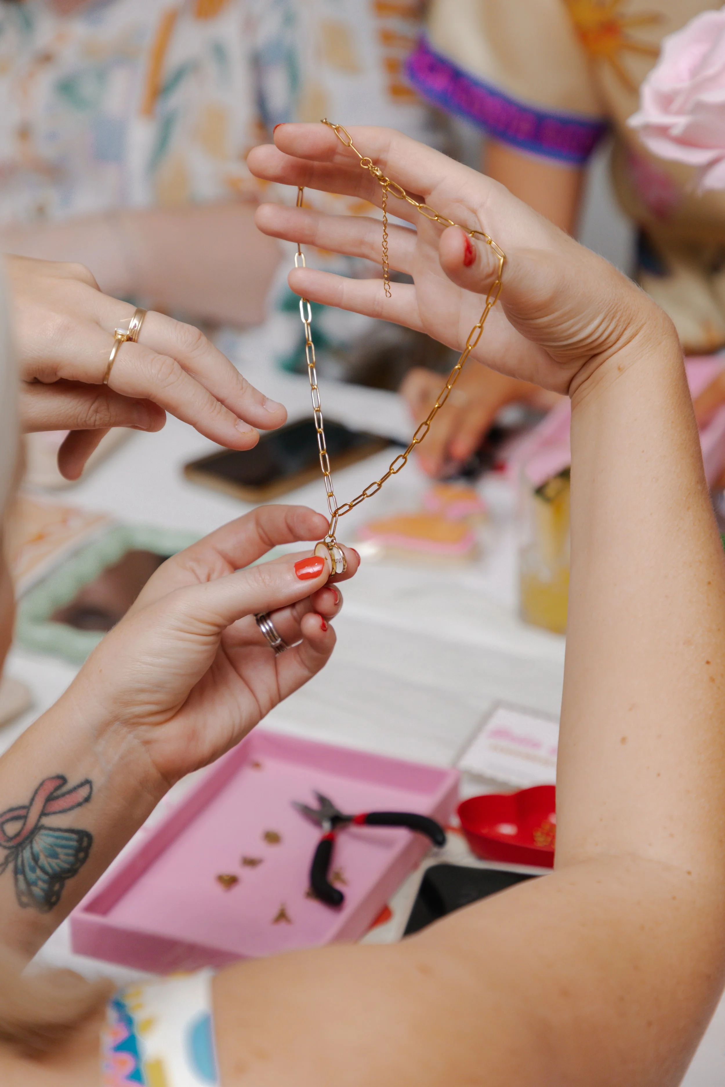 Two people exchanging a gold chain necklace at a jewelry table with pink jewelry box and jewelry-making tools in the background.