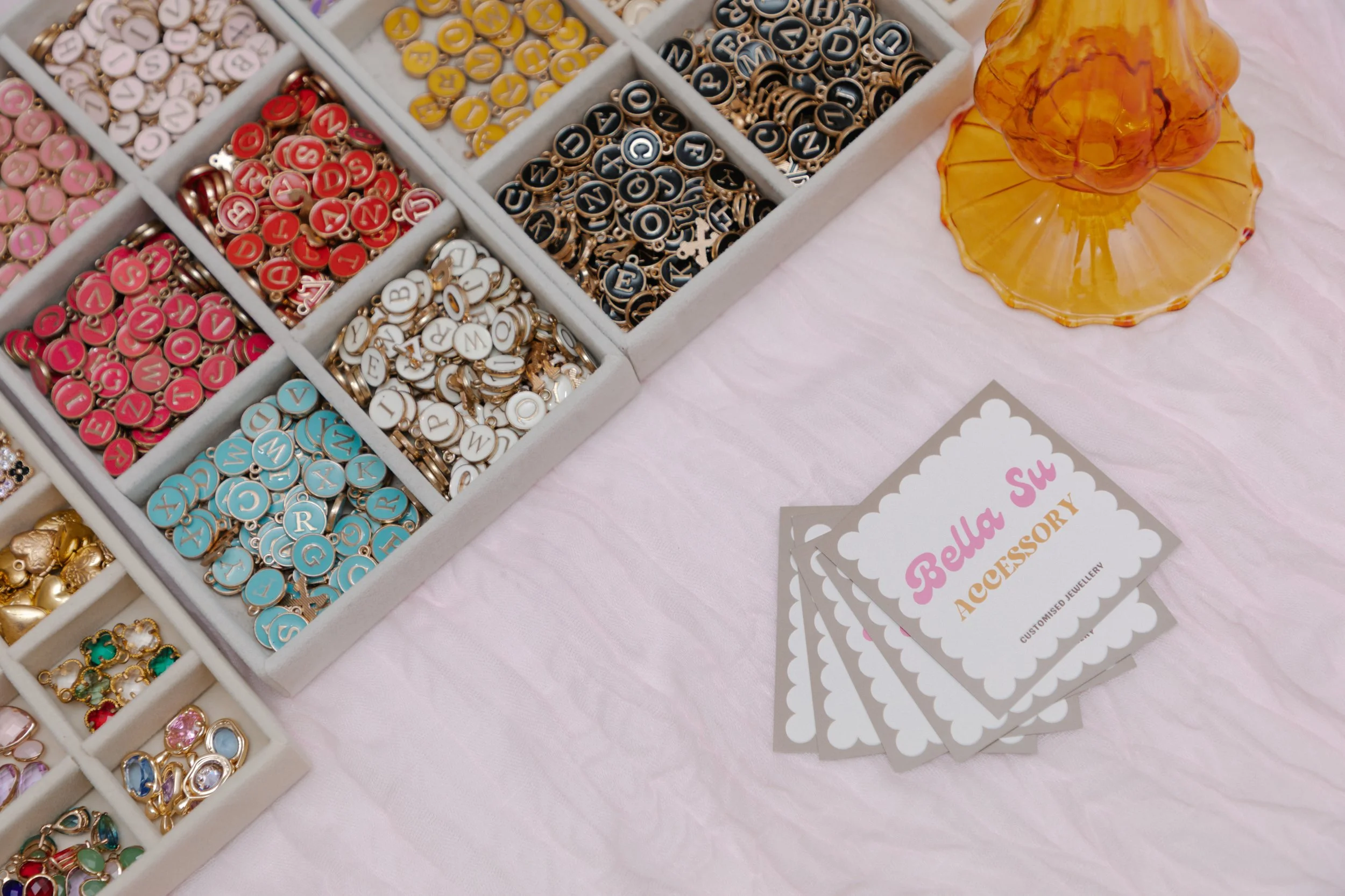 Display of colorful letter charms and jewelry accessories on a pink table, with business cards reading 'Bella Su Accessory'.