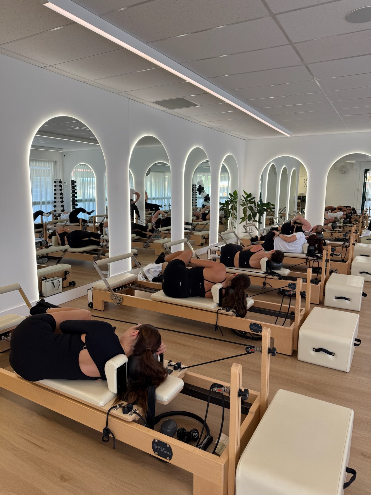 People practicing Pilates or physical therapy exercises on reformer machines in a bright, modern studio with white walls and arched mirrors.