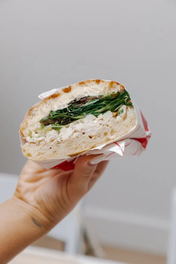 Close-up of a hand holding a sandwich with lettuce, chicken, and other fillings in a toasted bun, wrapped partially in red and white paper.
