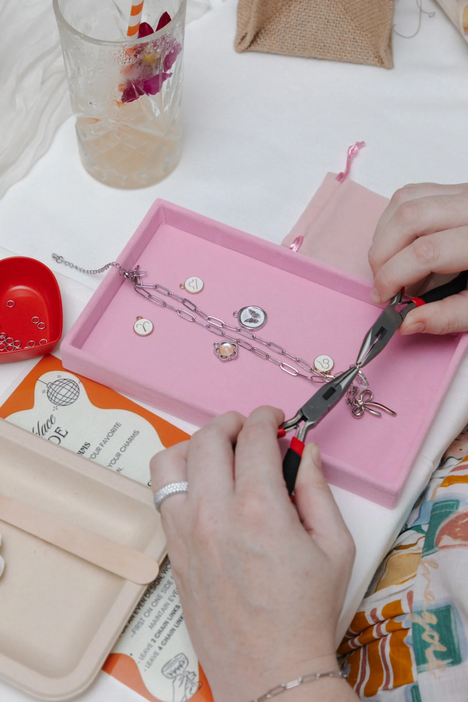Person repairing a bracelet using pliers on a pink jewelry box. Jewelry pieces are visible on the box, along with a glass of beverage and various small items on the table.