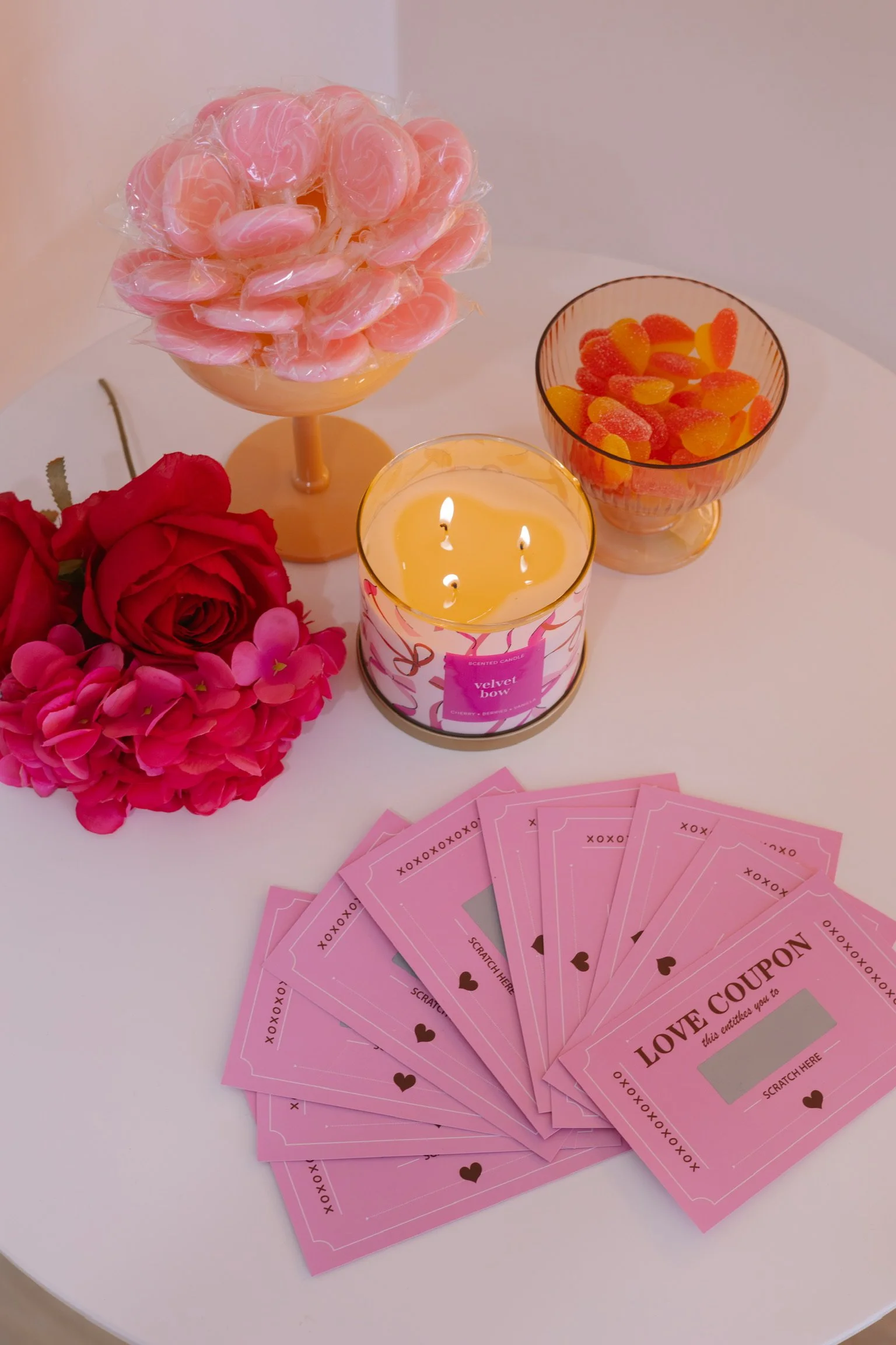 Valentine's Day gift setup with pink love coupons, pink candies, a candle, pink and red roses, and a bowl of gummy candies on a white table.