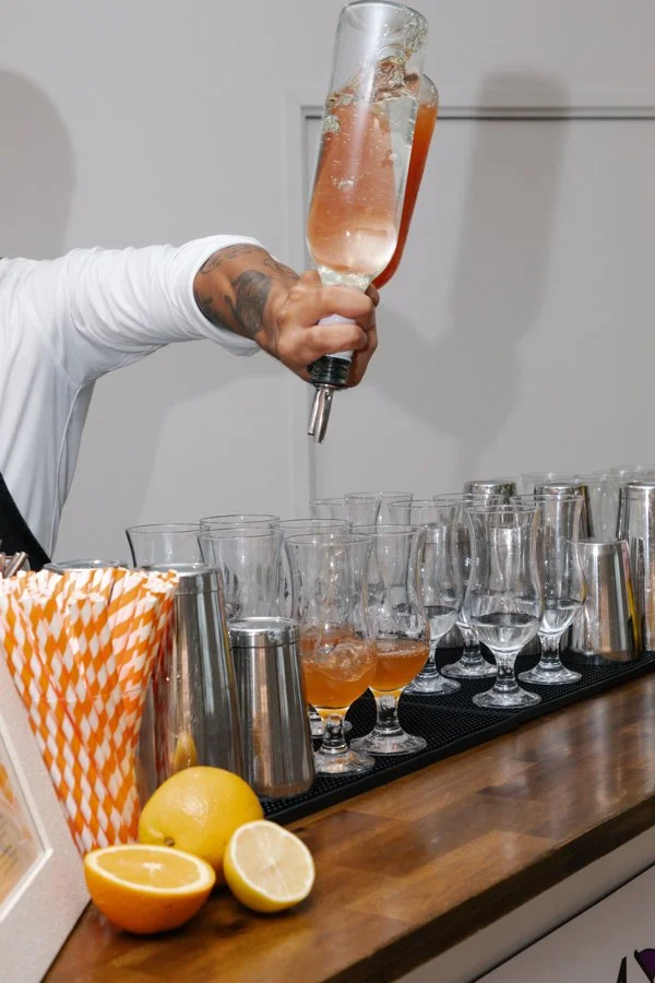 Person pouring a pink-colored cocktail or beverage from a bottle into cocktail glasses on a bar counter, with lemons and straws nearby.