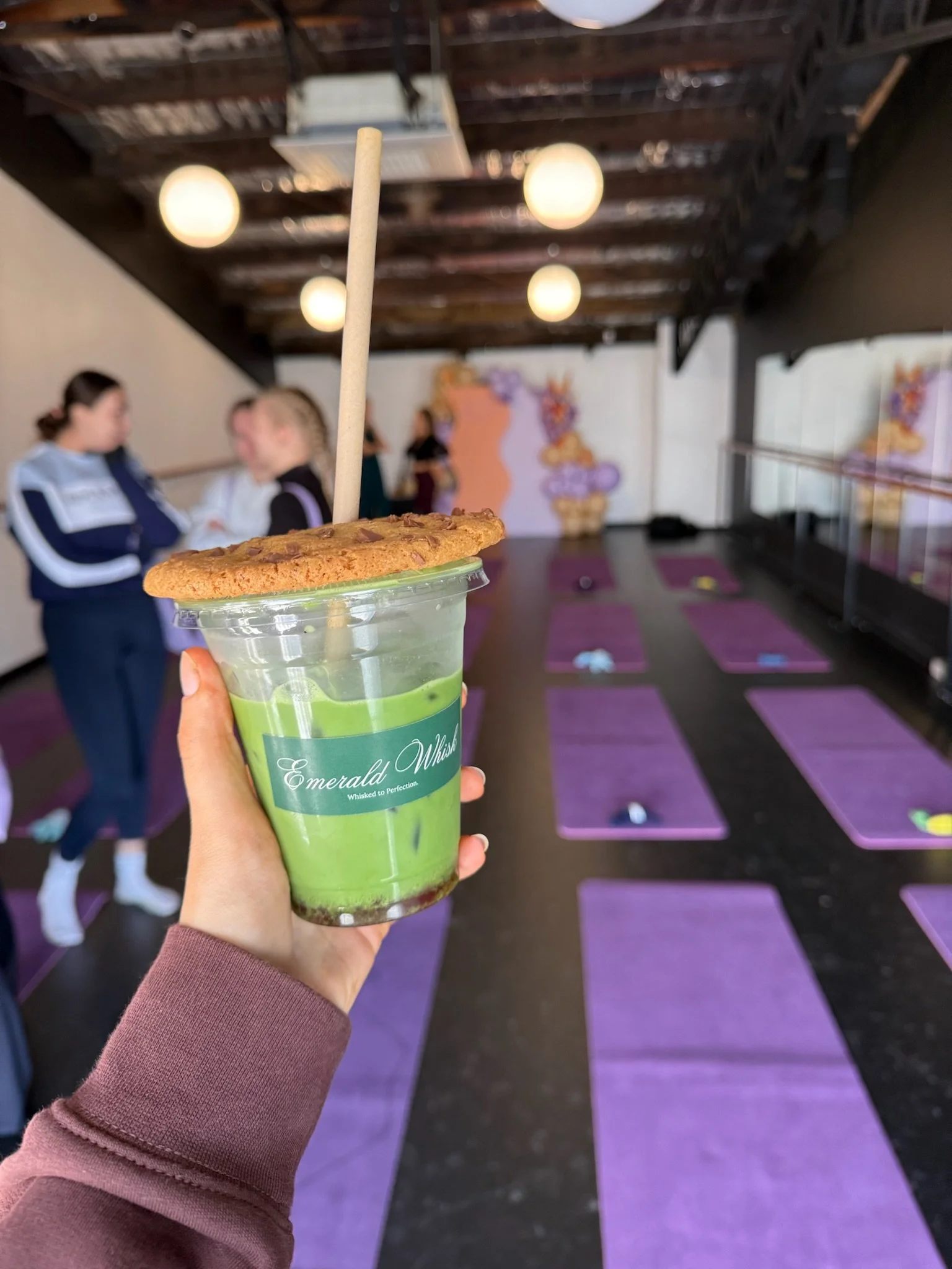 Hand holding a green matcha latte with a cookie on top and a straw, in a cafe with purple yoga mats on the floor and two women in the background.