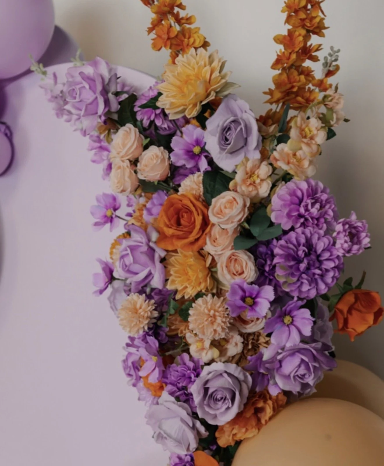 A floral arrangement of purple, orange, peach, and yellow flowers on a light purple table with purple balloons.