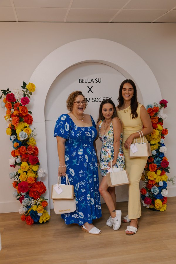 Three women standing in front of a white floral arch with the words 'Bella Su X SOM Society' on it, holding shopping bags, smiling at a floral event or photoshoot.