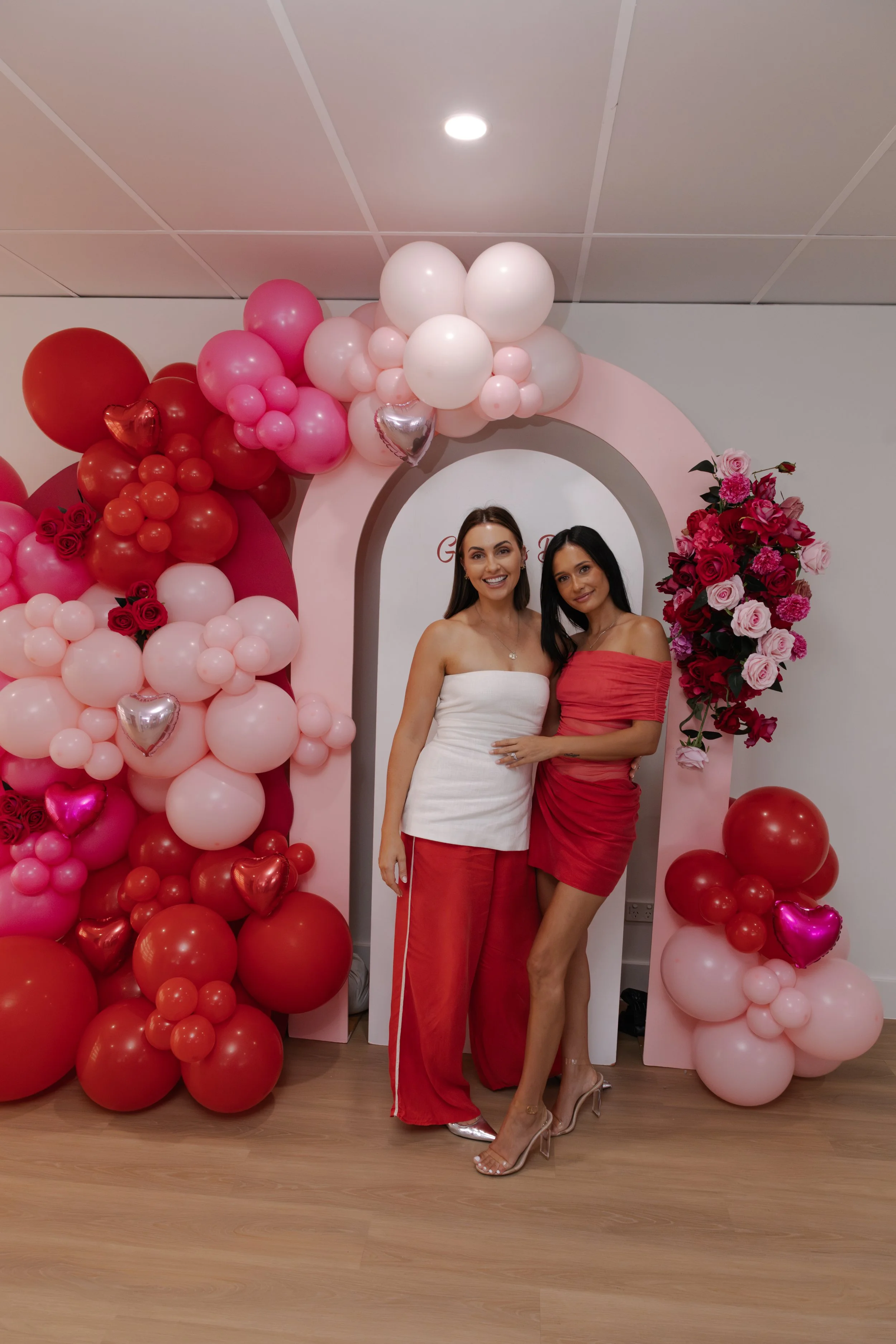Two women standing in front of a pink balloon arch with pink, red, and white balloons and flowers during a celebration or event.