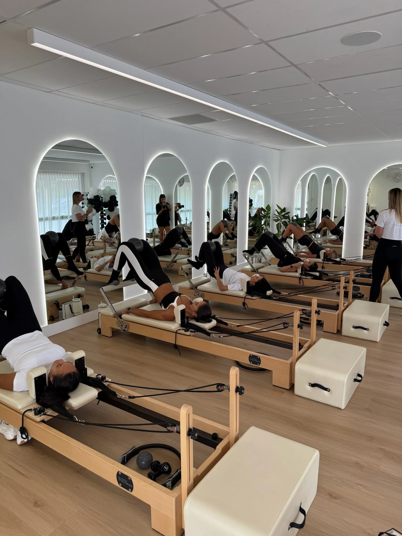 A pilates or fitness class in a bright, modern studio with women on reformer machines performing exercises, mirrored wall with LED lighting, and a presenter instructing.