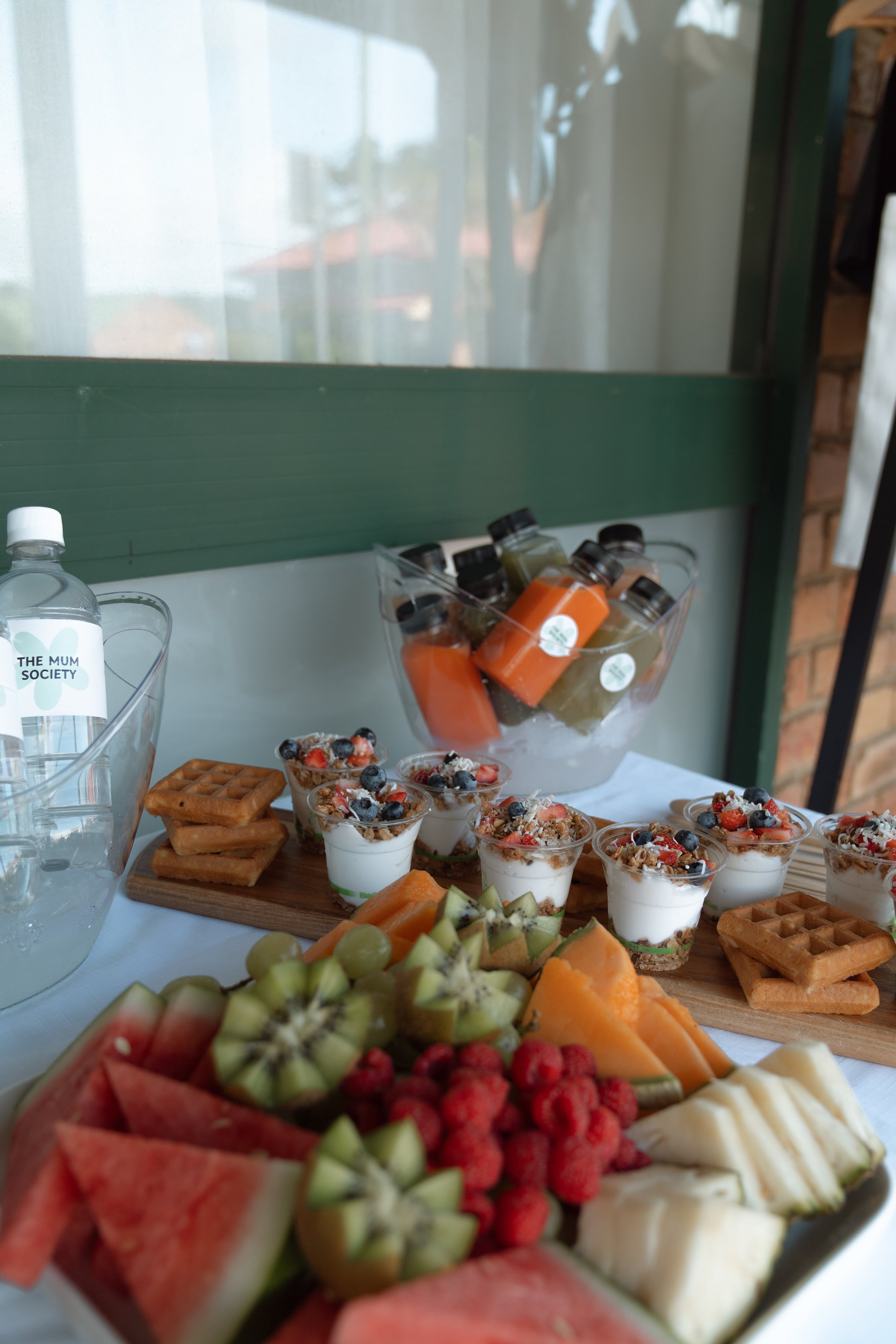 Assorted fresh fruit, yogurt parfaits topped with berries, small waffles, and juice bottles on a table.