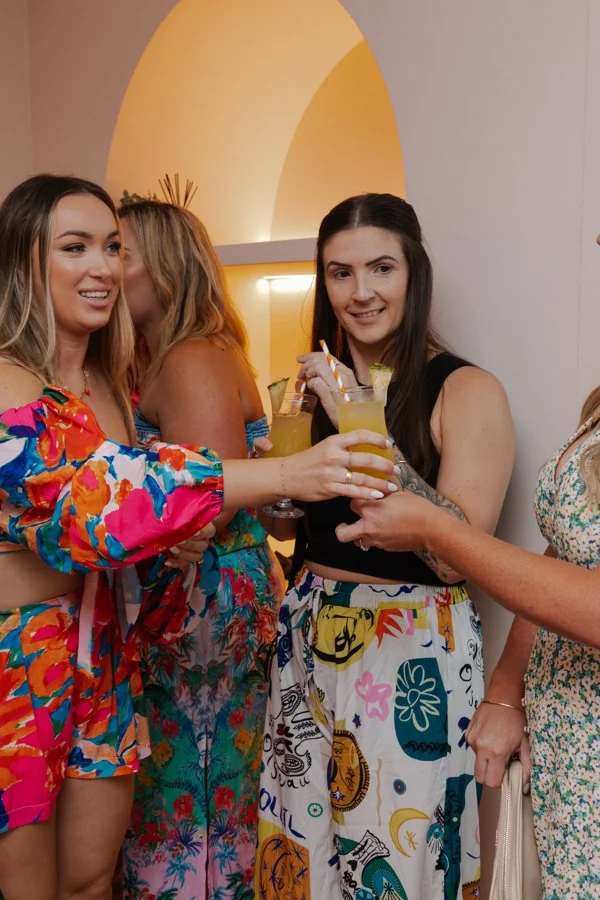 Group of women enjoying drinks at a social gathering or party, with one woman in a colorful floral outfit smiling and holding a drink, and others in vibrant clothing, standing indoors with warm lighting.