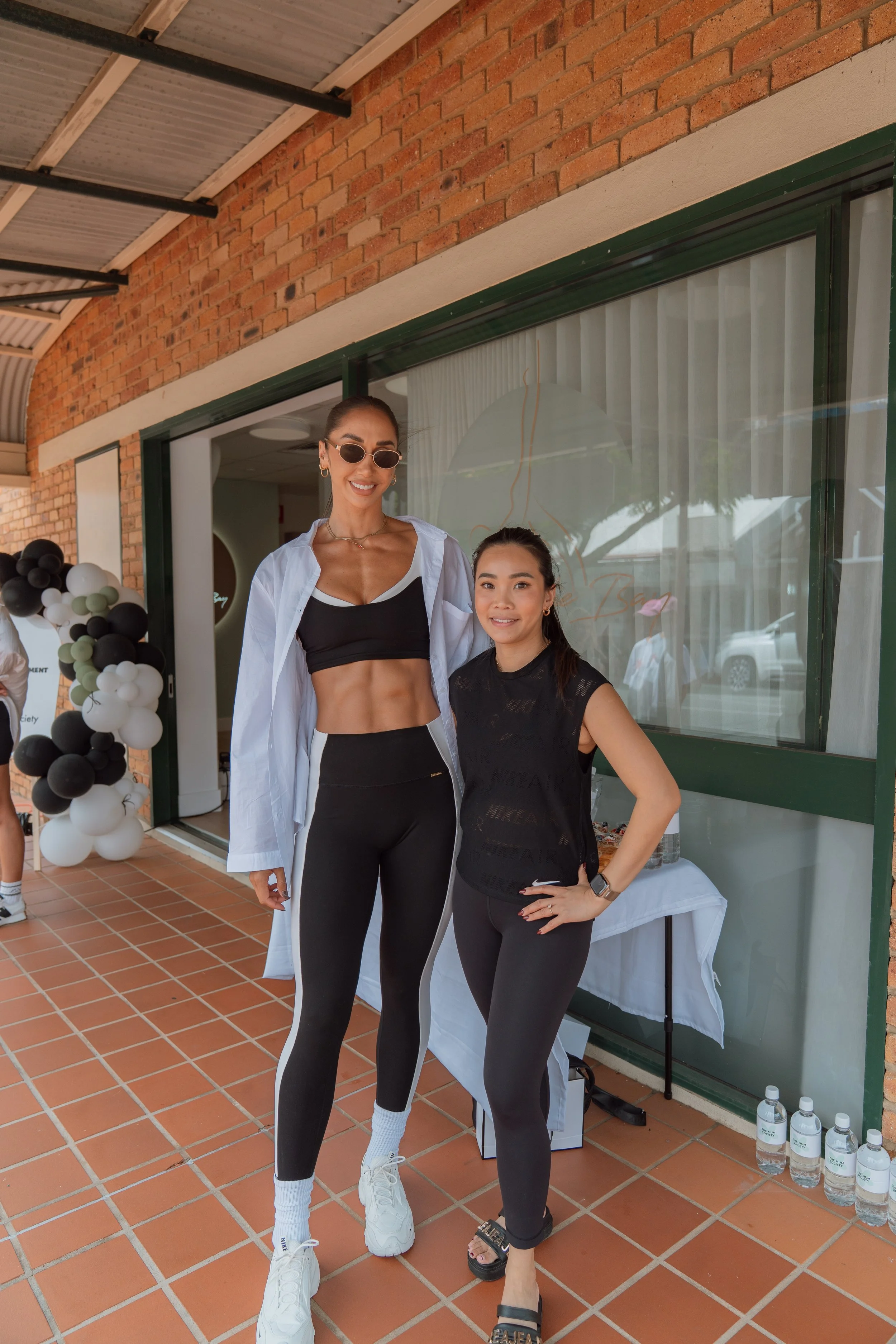 Two women posing outdoors in front of a brick building with large windows, one in black athletic wear and sunglasses, the other in black athletic clothing with a smartwatch, both smiling.