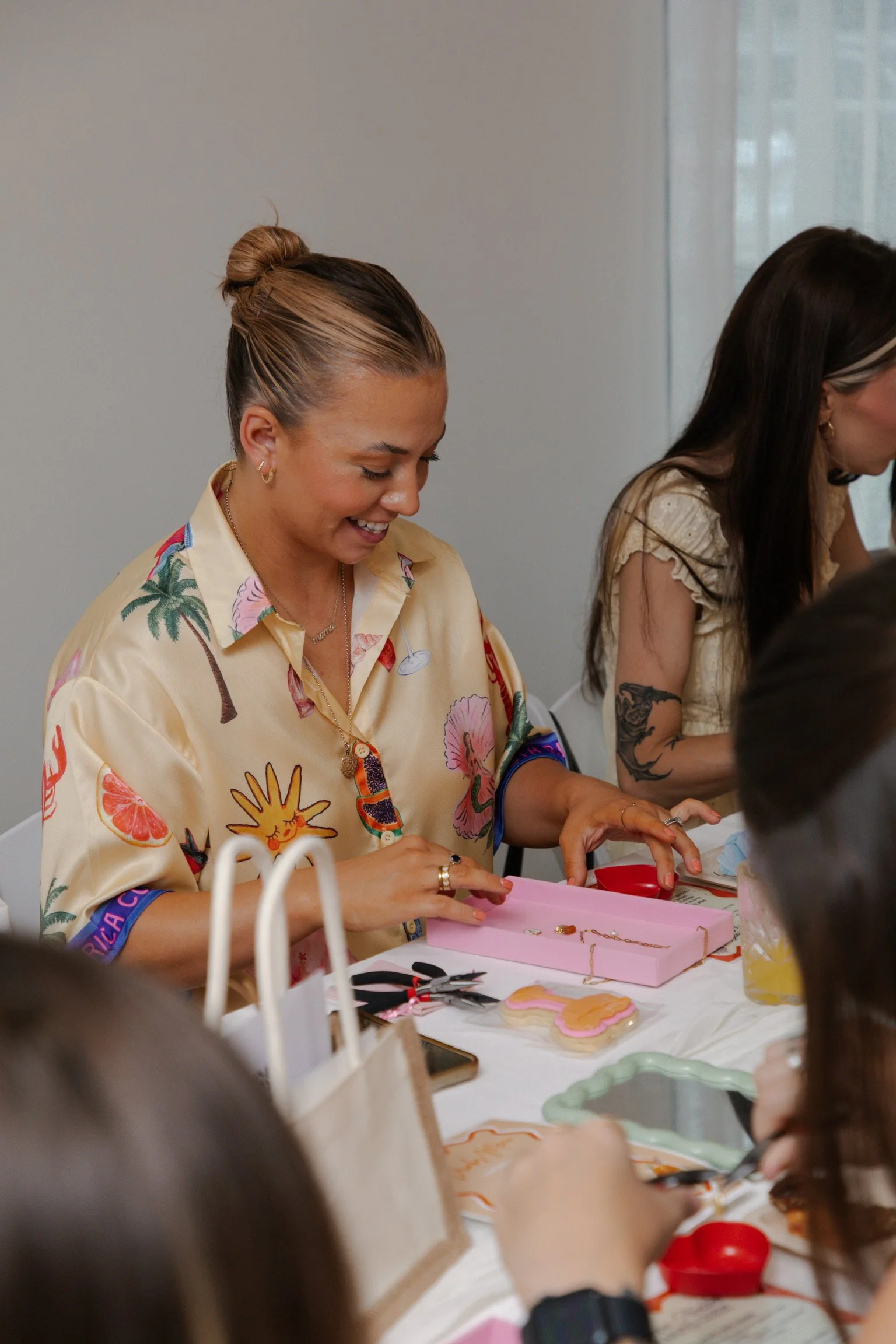 A woman with a bun hairstyle, wearing a colorful Hawaiian shirt, is smiling and organizing jewelry on a table.