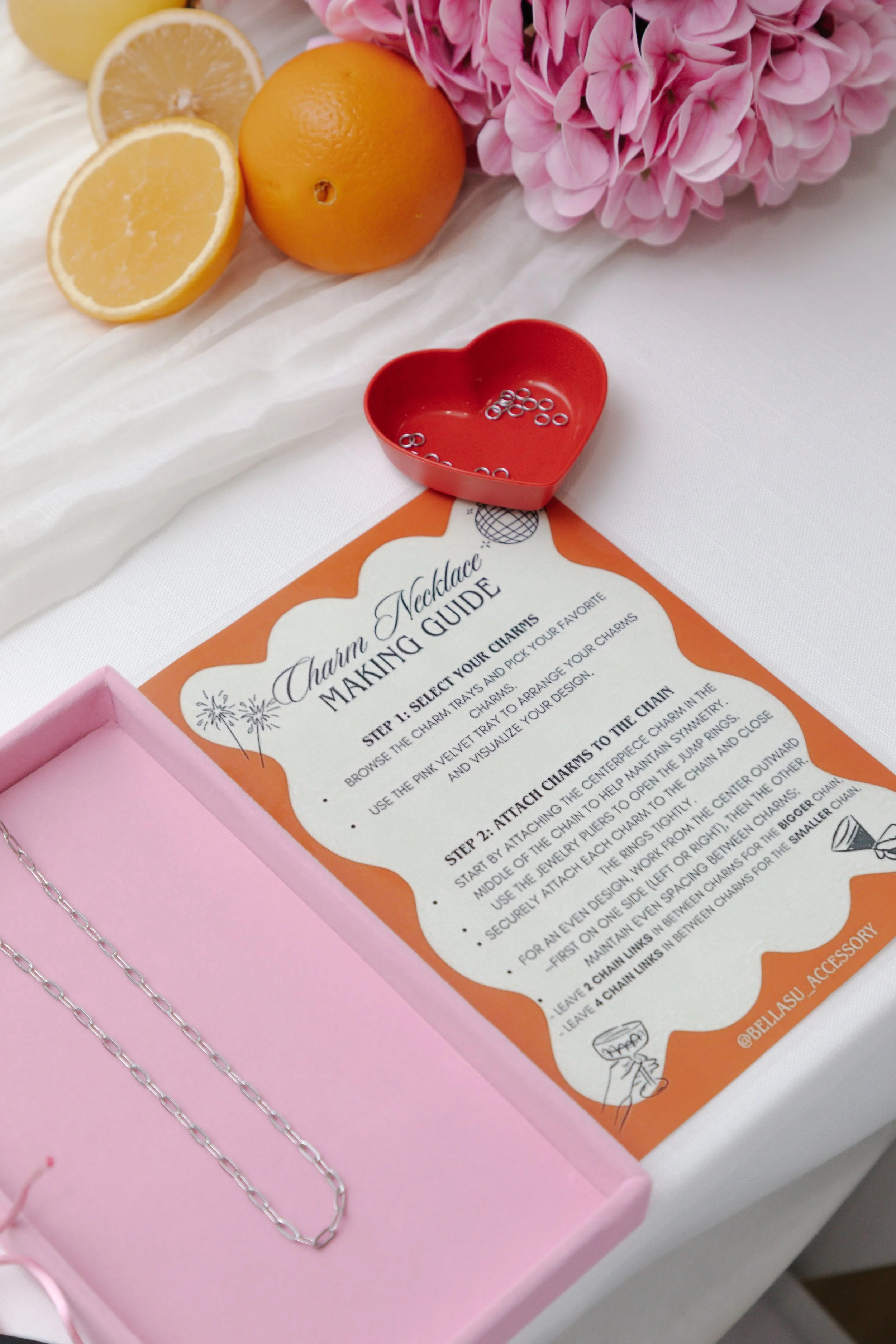 A charm necklace making guide, a small pink box with a necklace chain, a red heart-shaped dish with small rings, lemons, a pink hydrangea flower, and a white fabric surface.