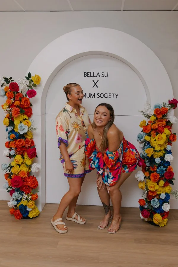 Two women in colorful, floral dresses standing in front of a white arch with a floral arrangement on each side. The arch has the words "BELLASU X AUM SOCIETY" written inside.