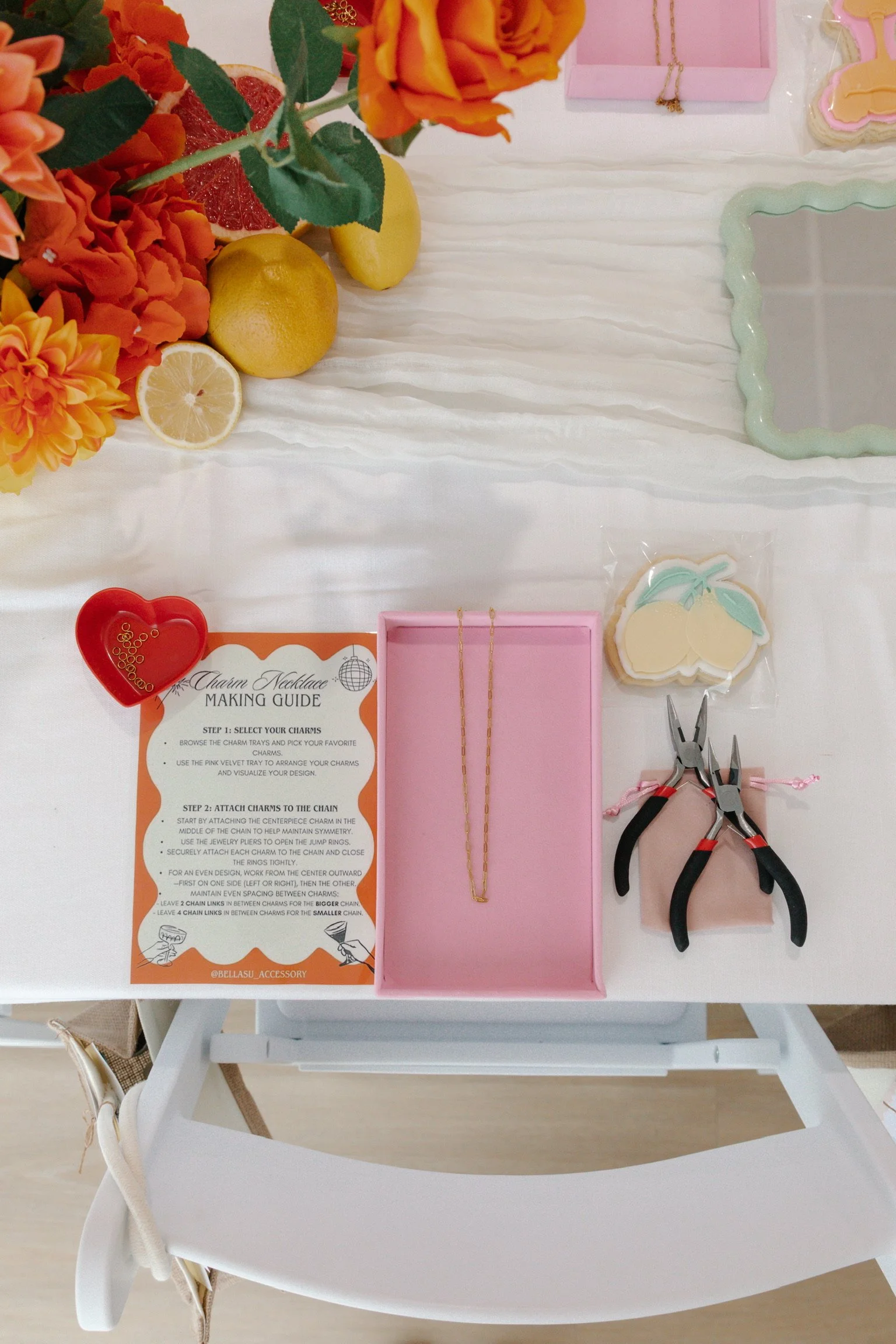 Girl's jewelry-making kit with charms, pliers, a necklace chain, and decorative cookies on a white table, with flowers and lemons nearby.