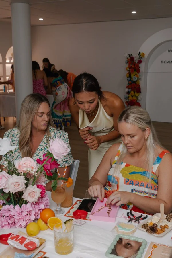 Women sitting at a table with flowers, snacks, and drinks, looking at a pink gift box, with another woman leaning over to look.