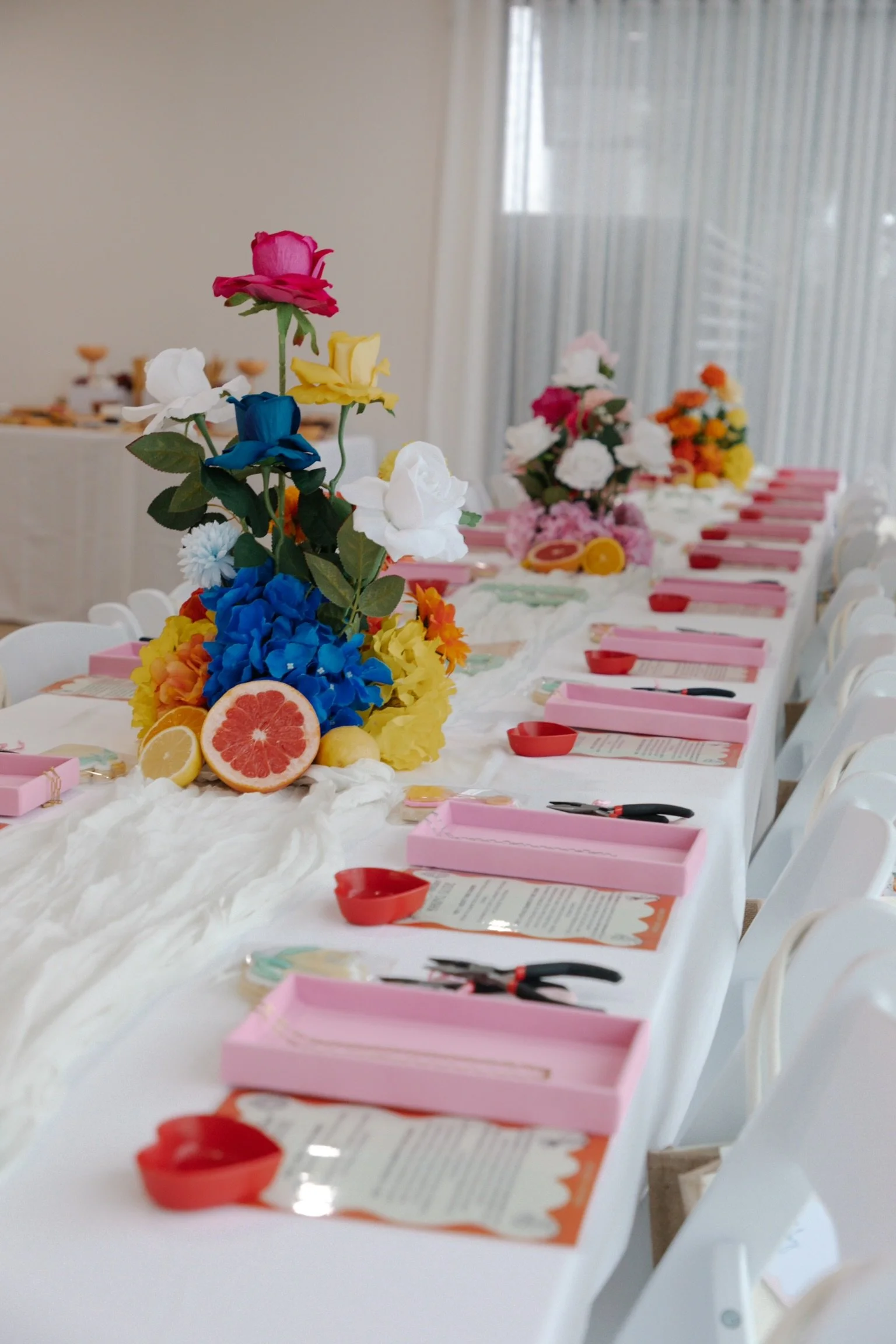 A long table set for a celebration with colorful floral centerpieces, pink boxes, printed menus, scissors, and small red bowls, with a window and curtains in the background.