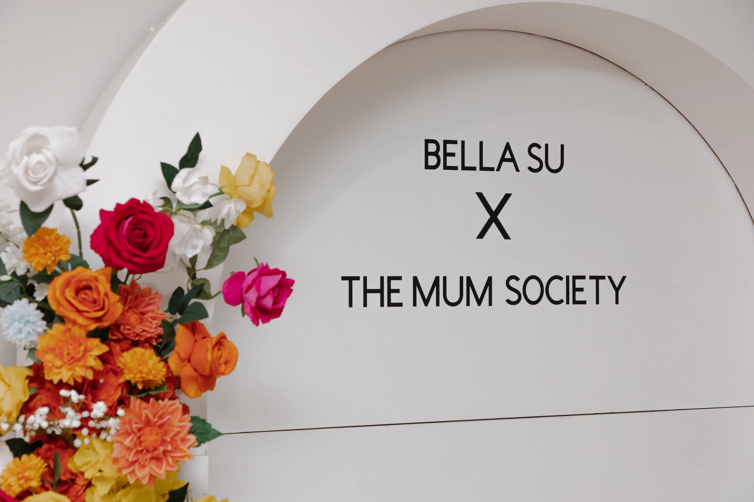 A white background with colorful flowers and black text that reads 'BELLA SU X THE MUM SOCIETY'.