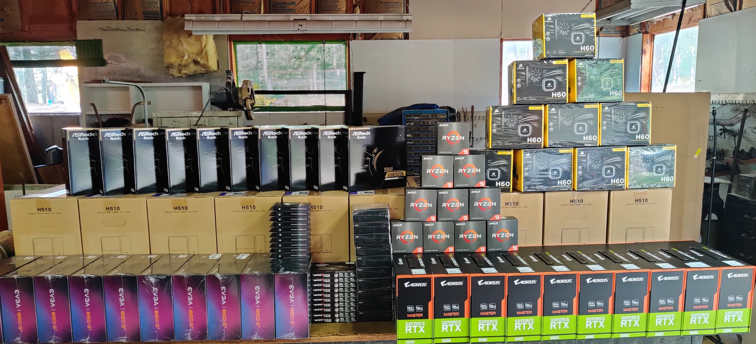 A large collection of computer hardware components on a workshop table, including multiple Nvidia GeForce RTX 3080 graphics cards in green and black boxes, AMD Ryzen 9 processors in black boxes, Aorus master motherboards, EVGA power supplies, ASRock server and rack components, and assorted other computer parts.