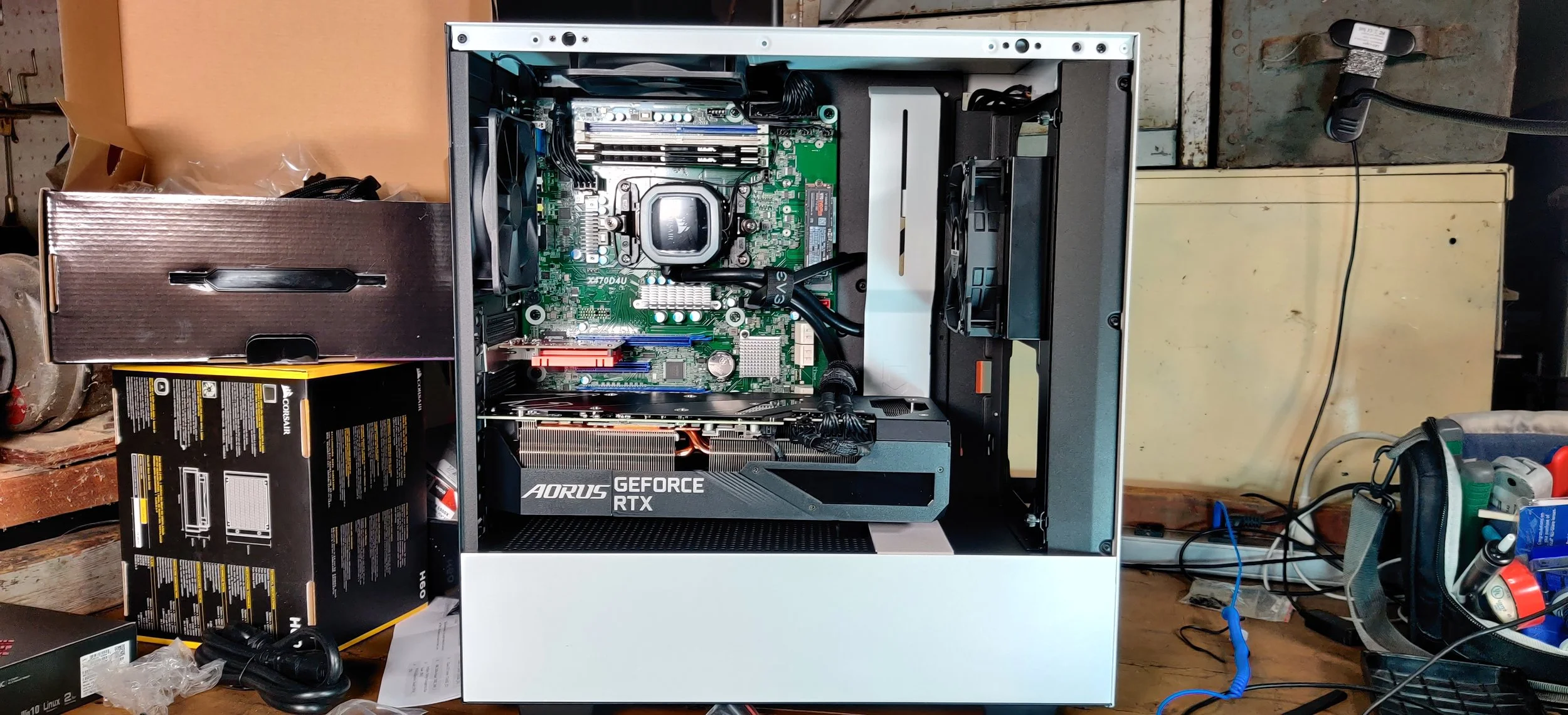 Open computer case with visible motherboard, graphics card, and cooling system, on a workbench with tools around.