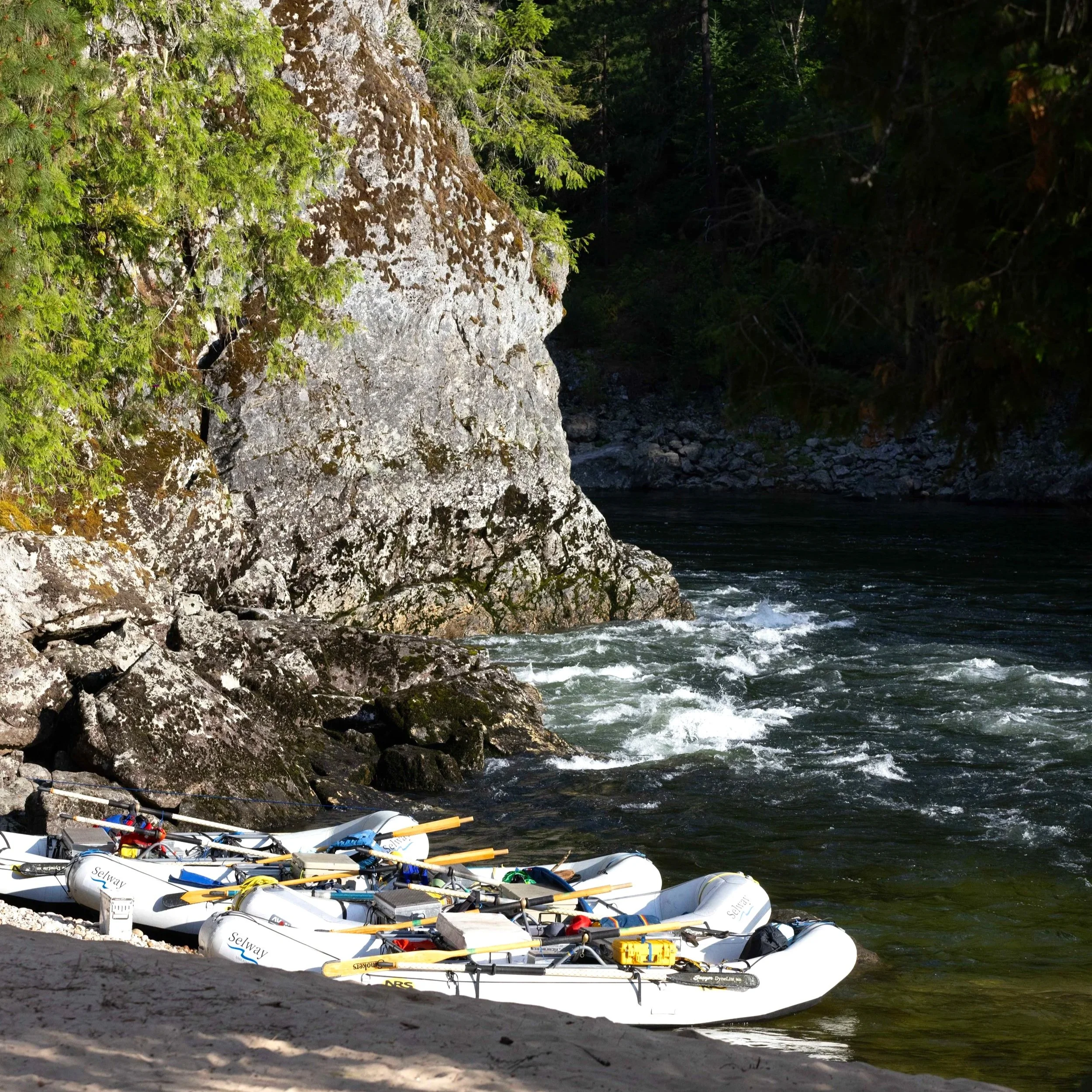 Four white rafts along the sandy river's edge with a large rock wall in the background lit by sunlight. Small white waves on the river are also in the background.