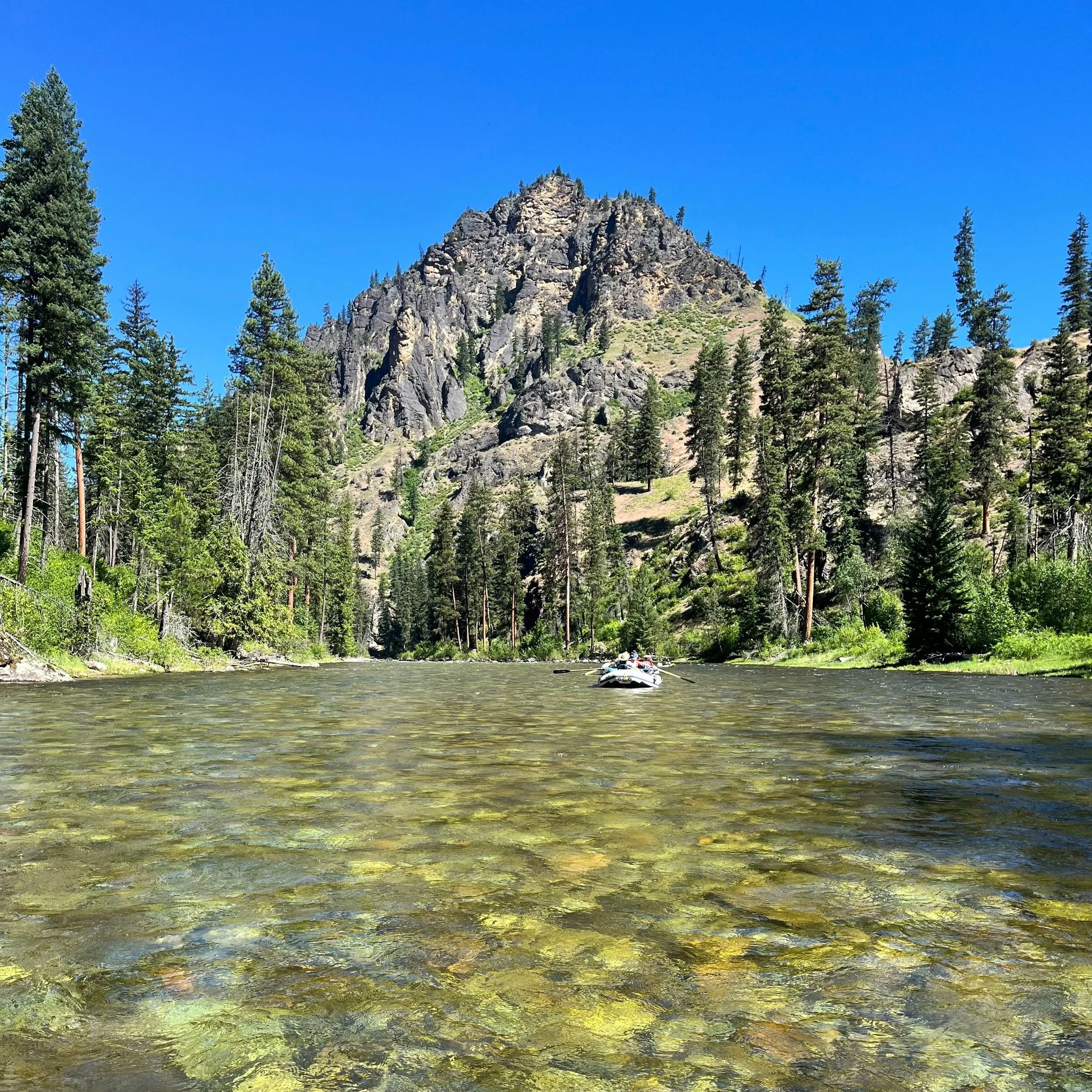 One white raft in the distance, floating down a crystal clear river toward a craggy mountain peak. The sun is shining through the water showing the rocks below.