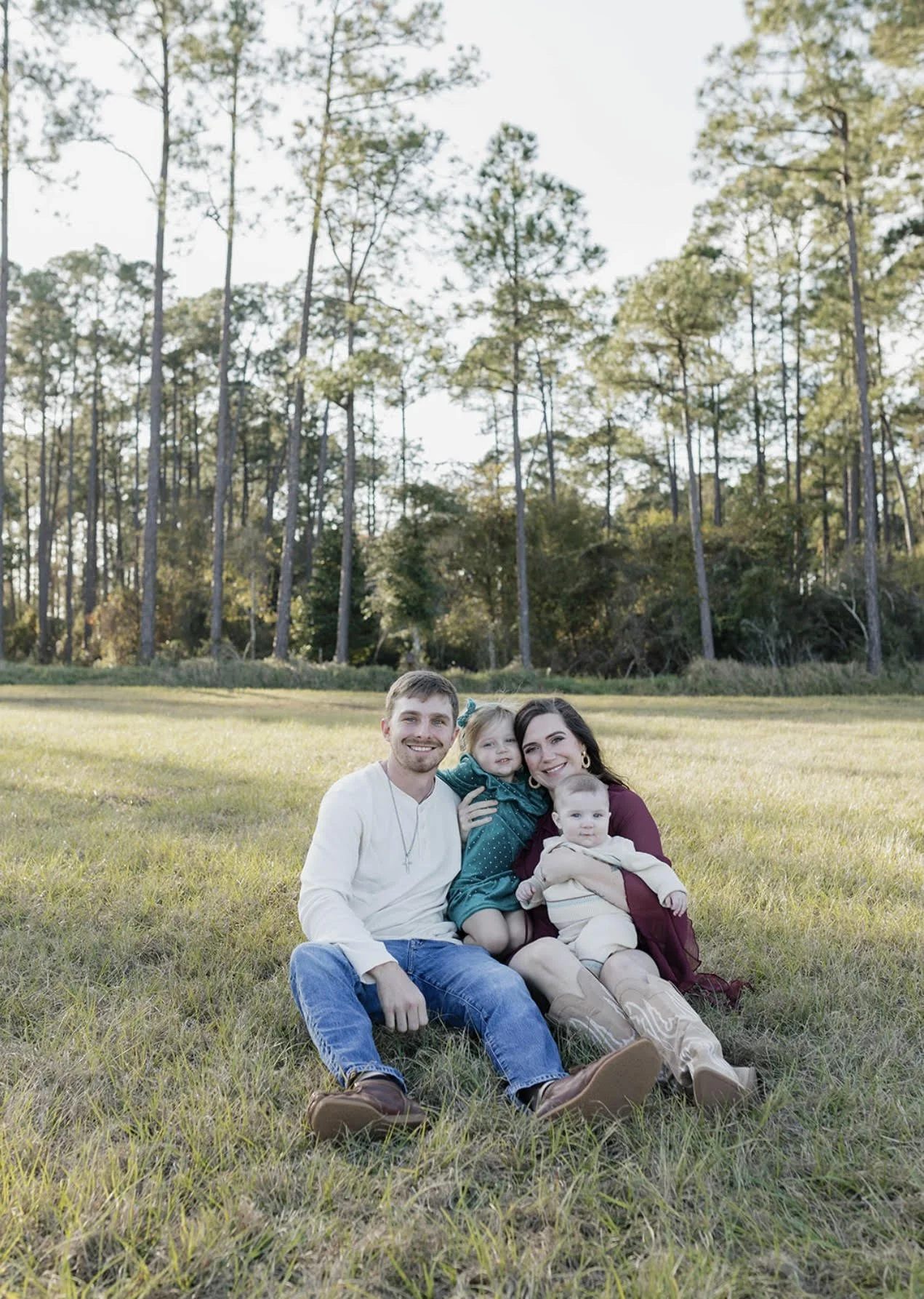 A happy family of four sitting together on the grass in a field with tall trees in the background.
