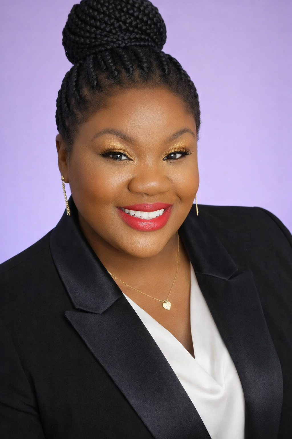 Portrait of an African American woman with braided hair styled in a high bun, wearing a black blazer over a white top, gold earrings, a gold heart necklace, and red lipstick, smiling against a purple background.