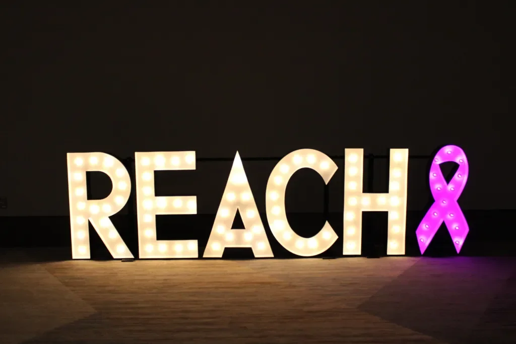 Large illuminated sign spelling 'REACH' with a pink awareness ribbon at the end.