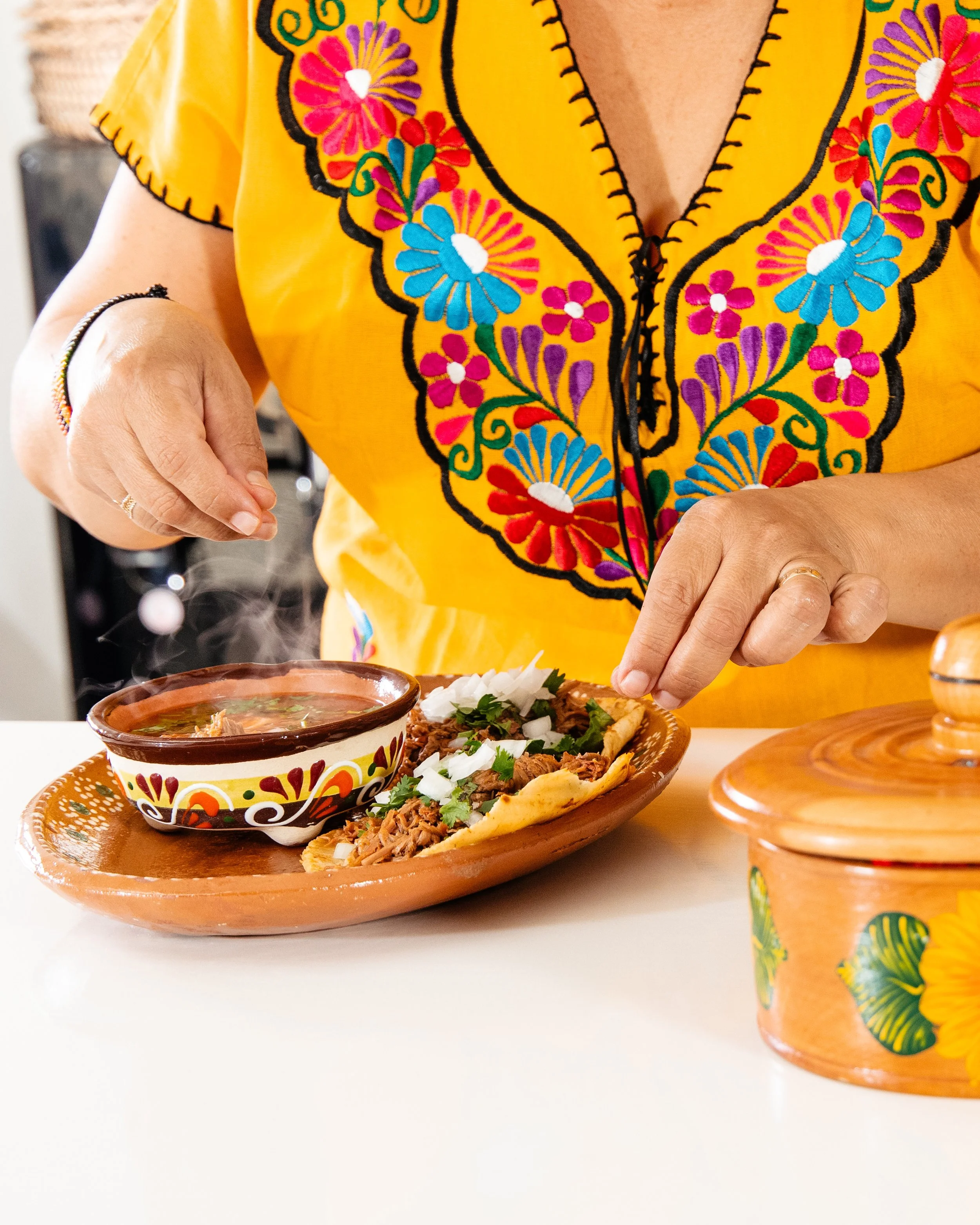 A person wearing a yellow embroidered blouse is preparing traditional Mexican food on a molcajete. The dish includes shredded meat, diced onions, and cilantro, alongside a bowl of steaming soup, all served on a decorated terracotta plate and surrounded by colorful Mexican ceramics.