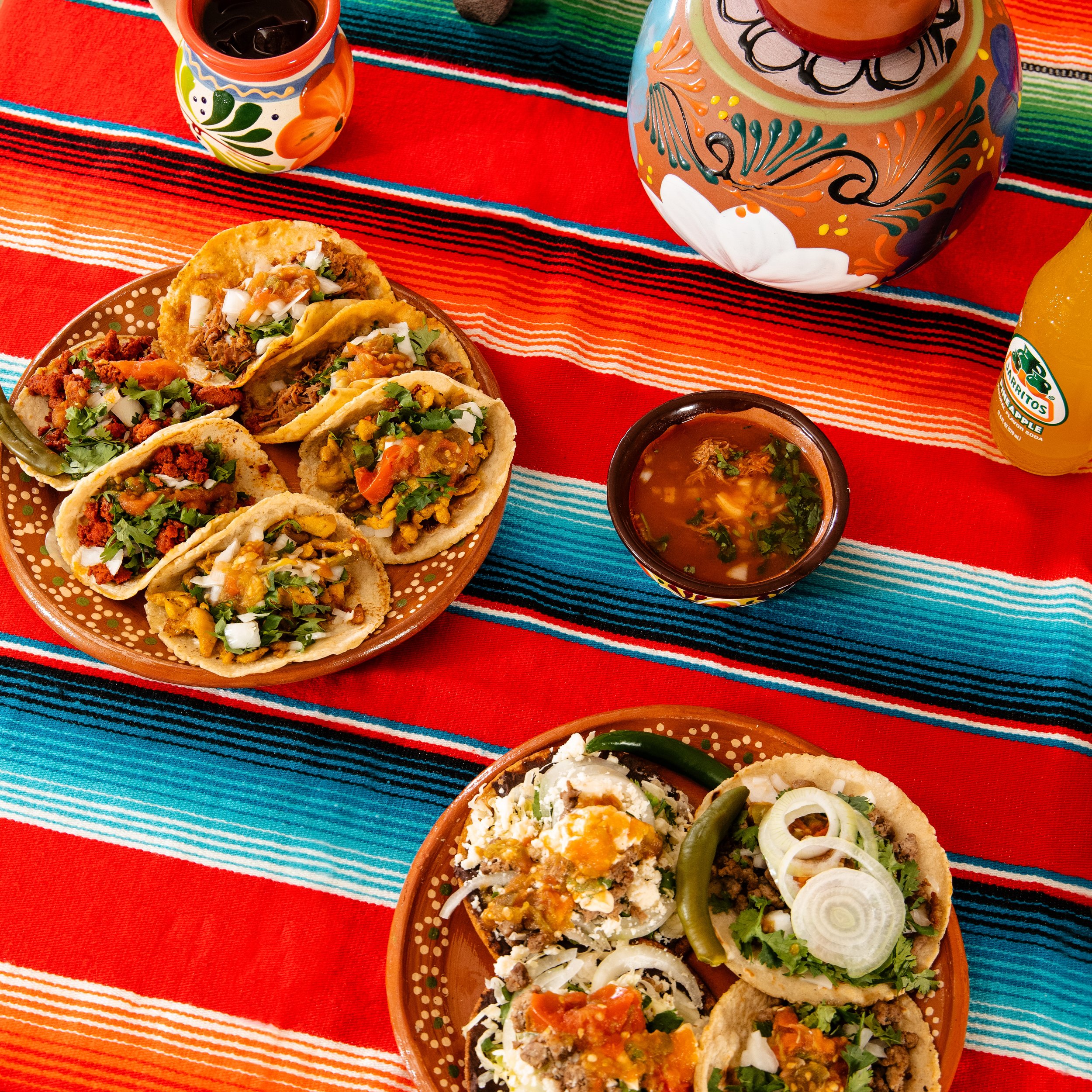 Assorted traditional Mexican tacos with various fillings, a bowl of soup, a bottle of Jarritos soda, and colorful Mexican tablecloth.