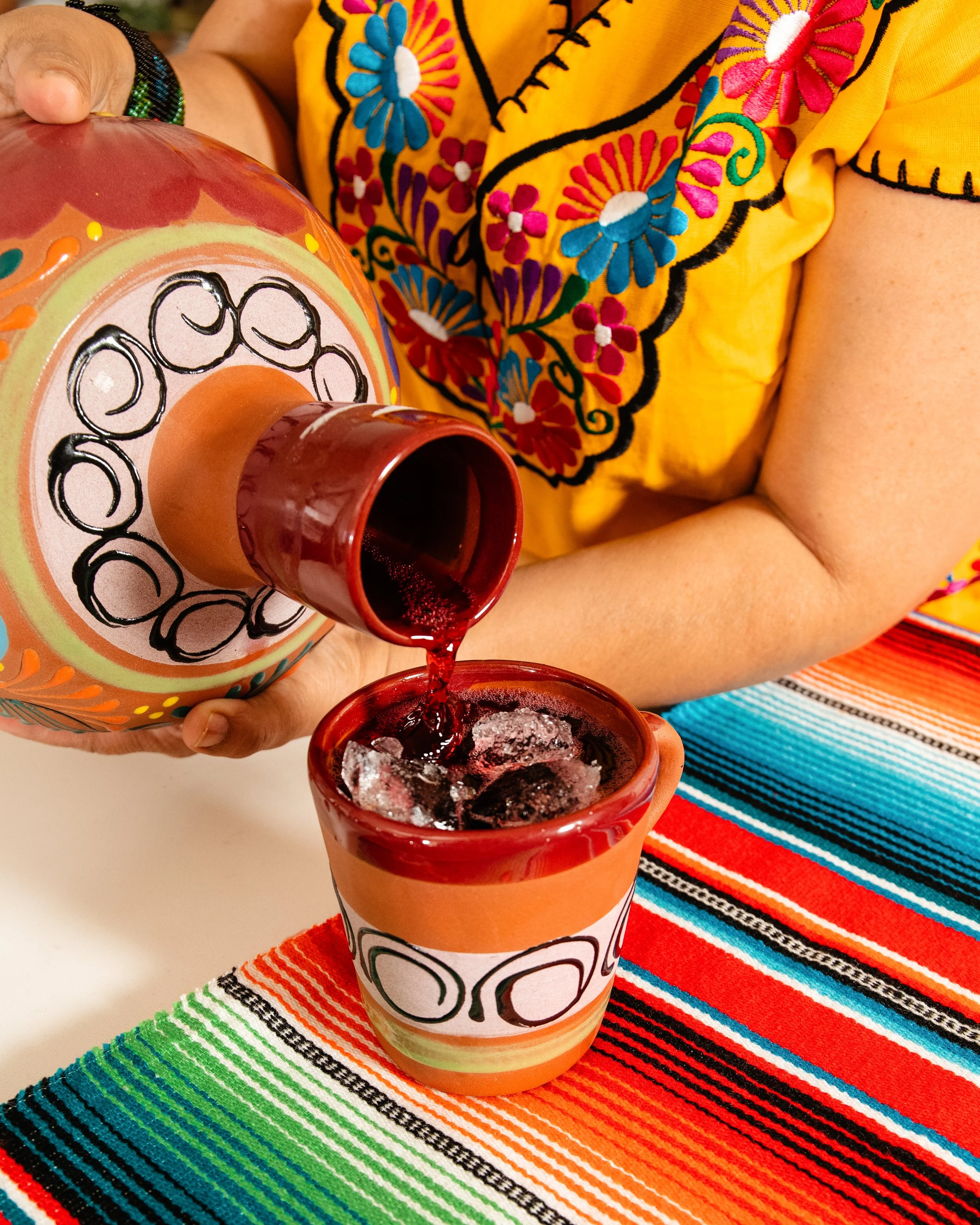 Person pouring red beverage from a decorative bottle into a matching glass on a colorful striped tablecloth.