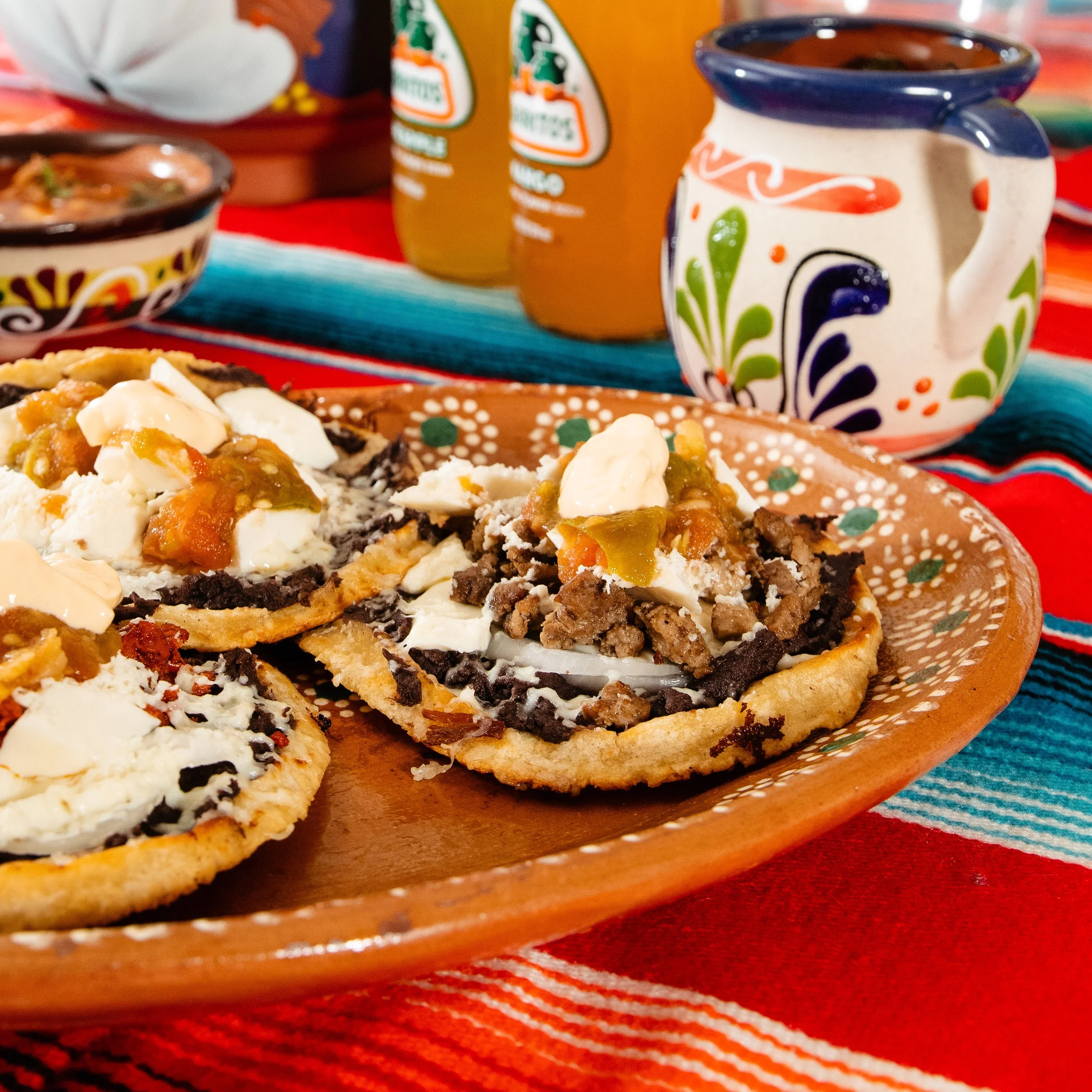 Close-up of a traditional Mexican dish served on a decorative plate, topped with cheese, and salsa, set on a colorful woven tablecloth with bottles of orange soda and a ceramic coffee mug in the background.