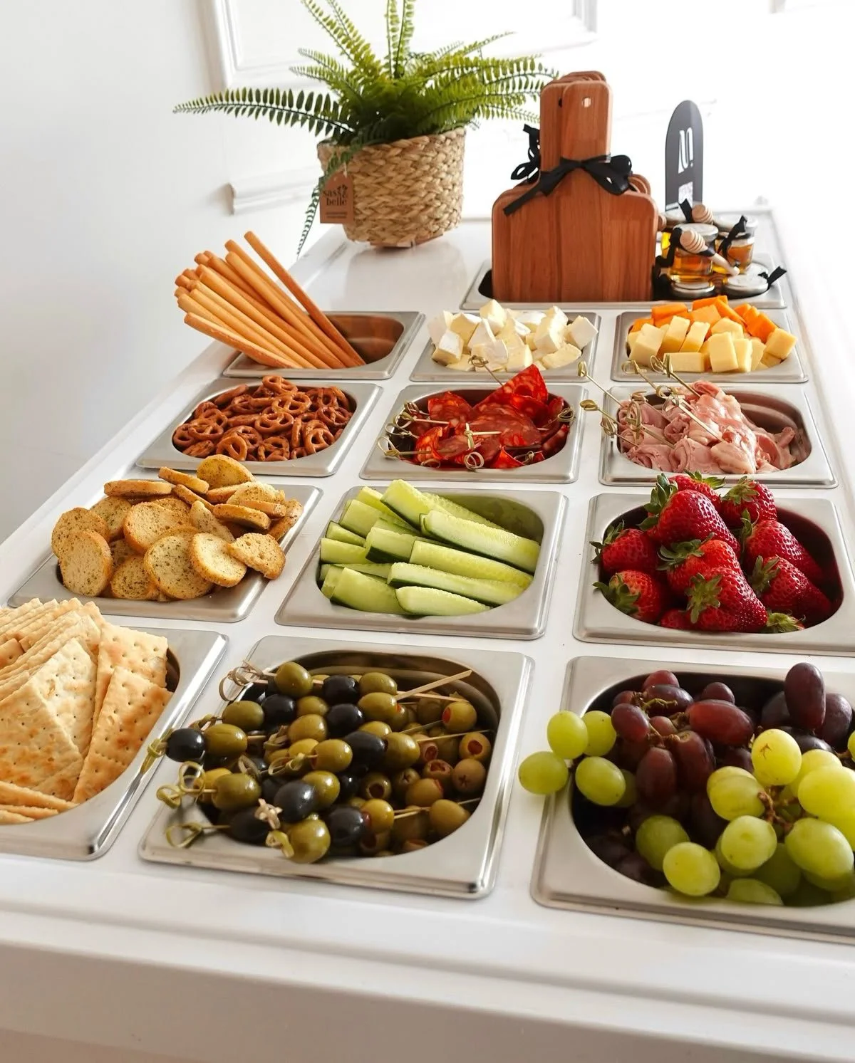 A breakfast or brunch buffet with various fresh fruits, vegetables, sliced meats, cheeses, crackers, bread, pretzels, breadsticks, and olives arranged in metal trays on a white counter.