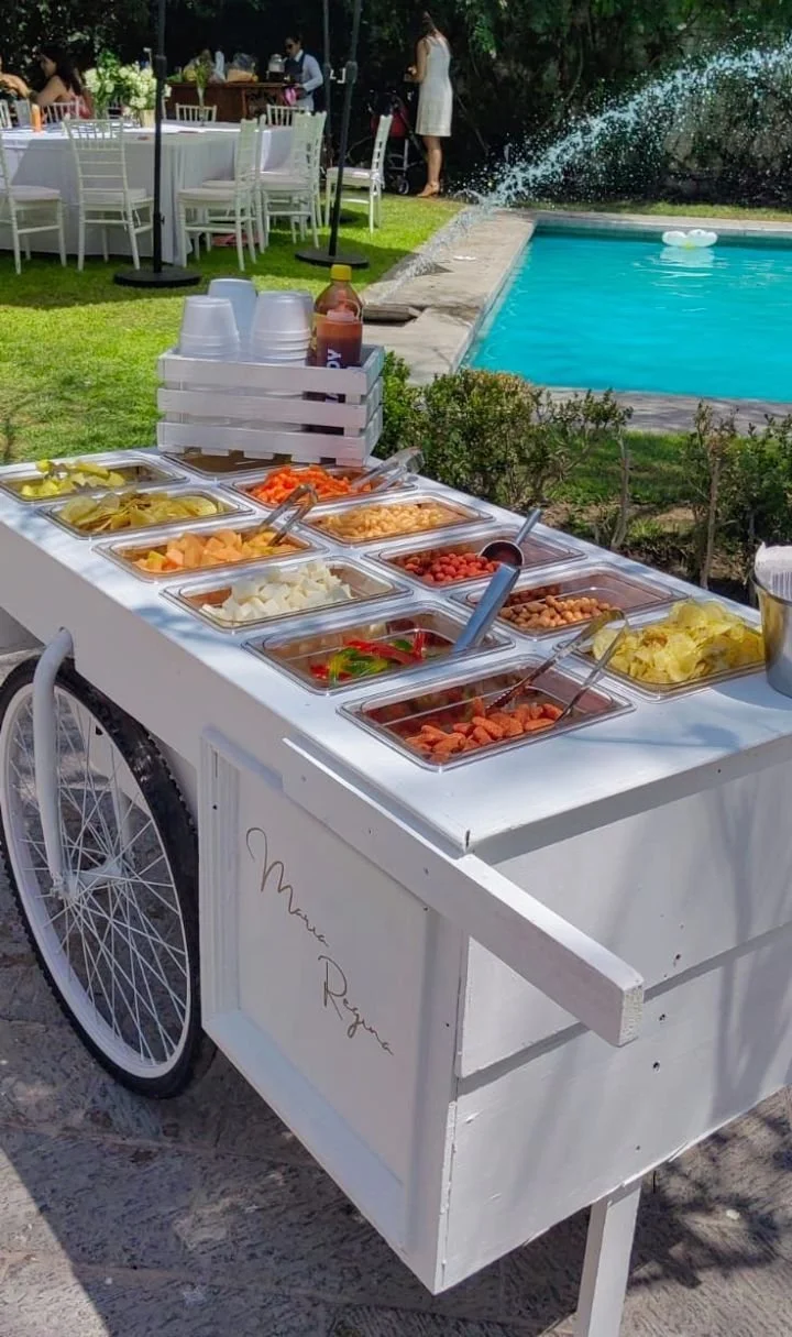 A white mobile buffet cart with various containers of chopped fruits and vegetables, positioned near a swimming pool at an outdoor gathering. In the background, there are tables with white chairs and a group of people, including a woman standing near the pool.