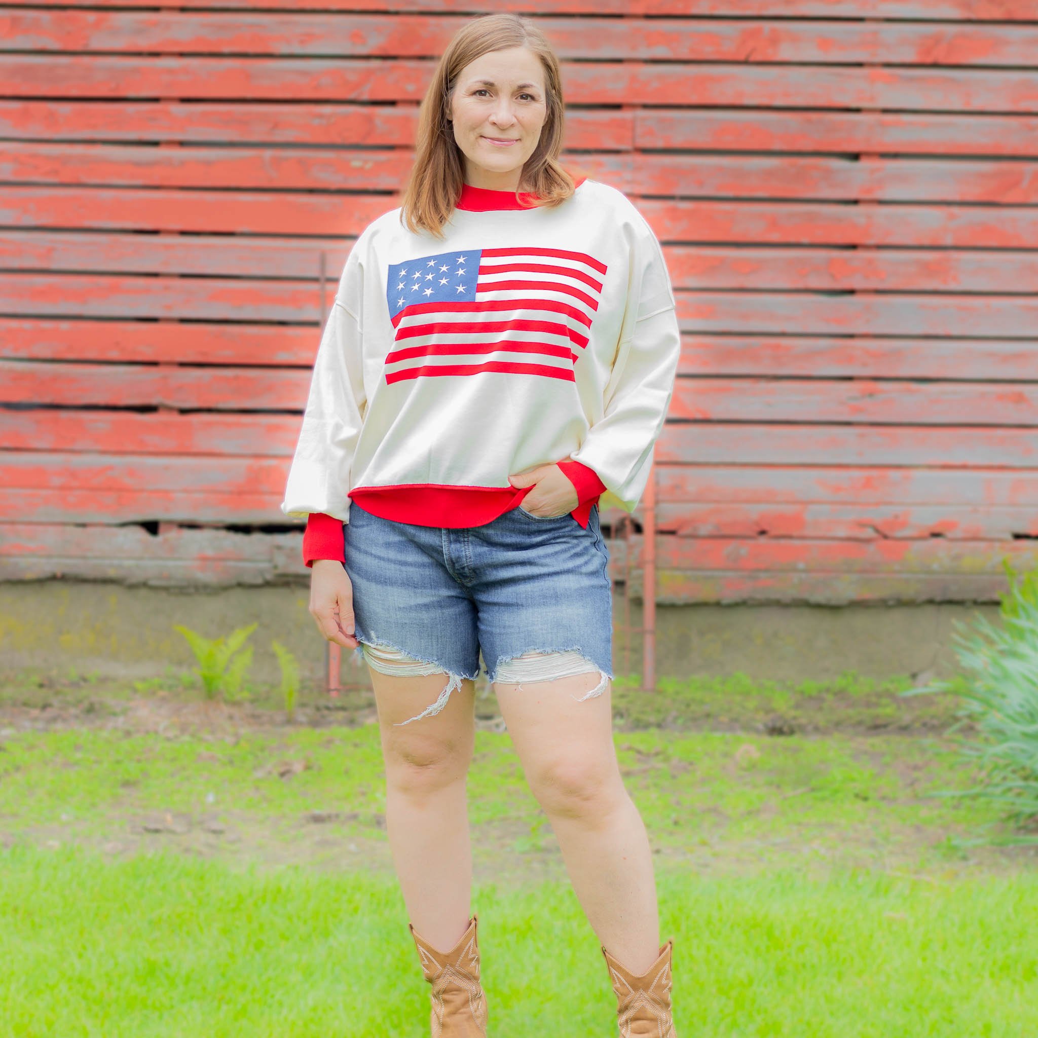 woman wearing white sweater with red trim and American flag across the chest, jean shorts and boots beside a barn.