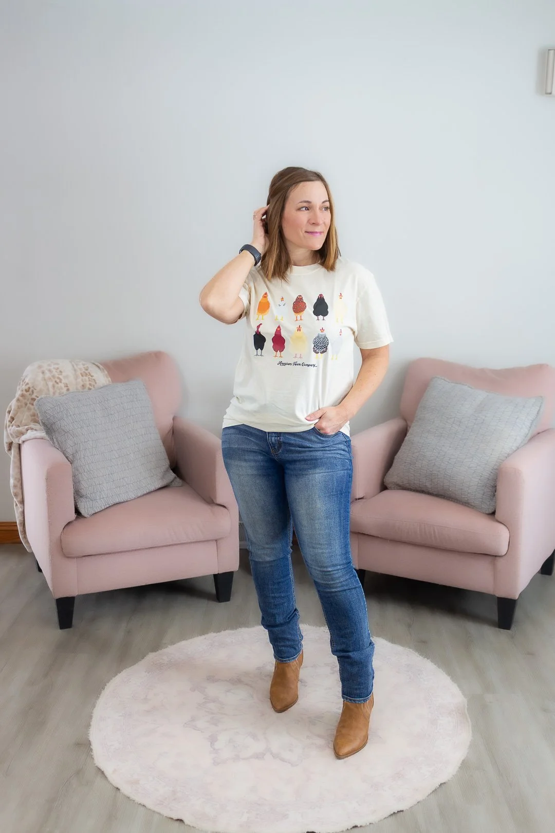 Woman standing on a pink rug in a living room, wearing a white T-shirt with colorful chickens, jeans, and brown boots, with two pink armchairs and gray pillows behind her.