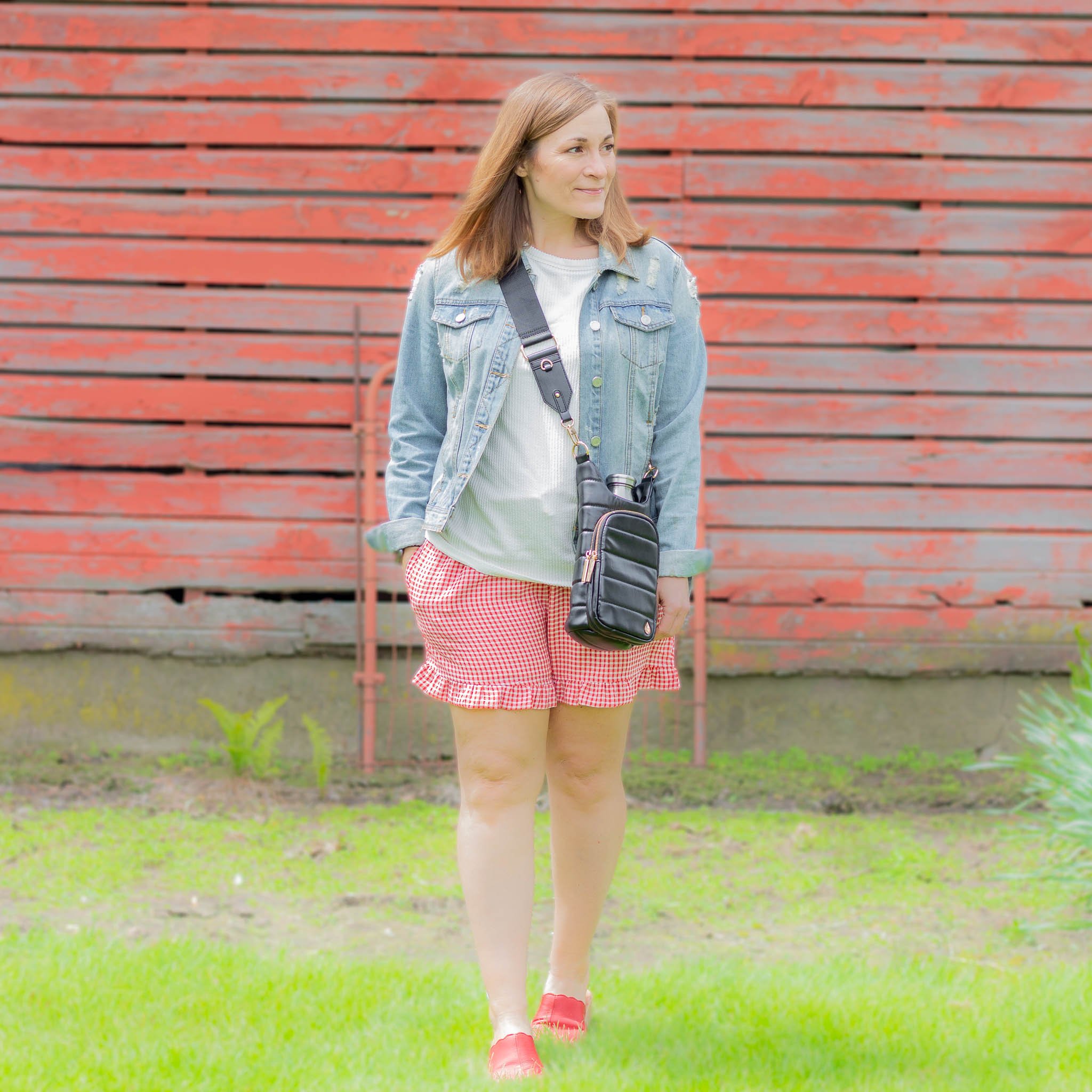 woman walking by barn wearing red gingham shorts, white tee, denim jacket and black crossbody bag.