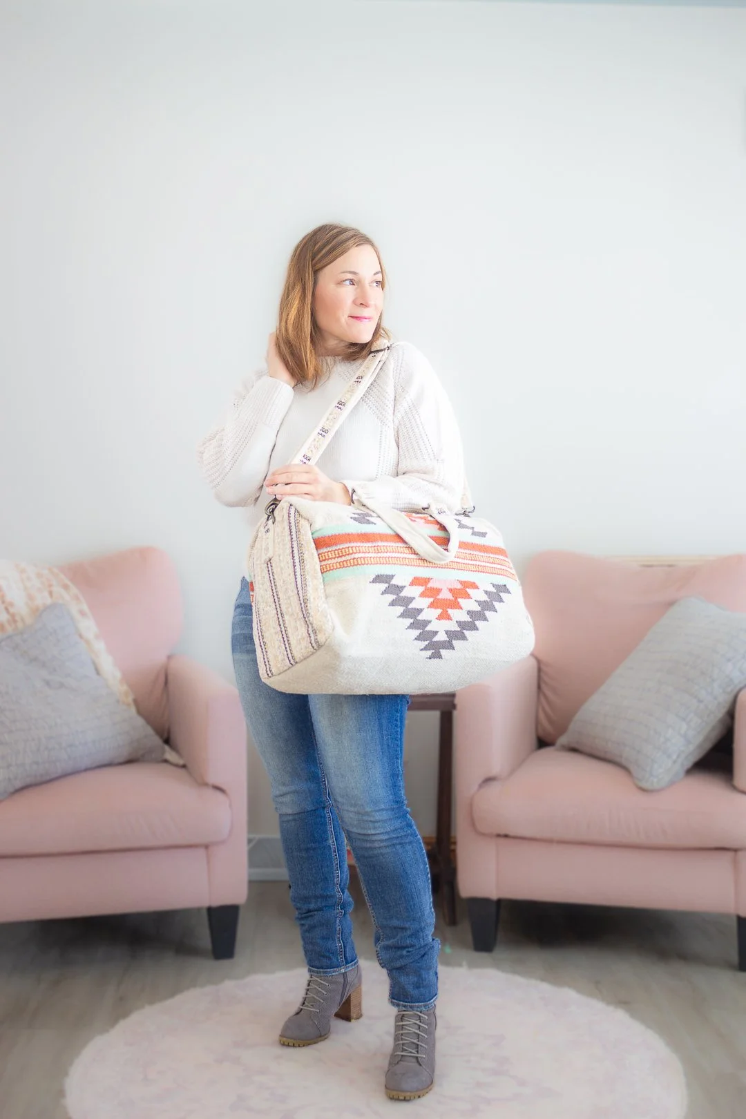 A woman holding a large patterned tote bag standing in a living room with pink couches and decorative pillows.