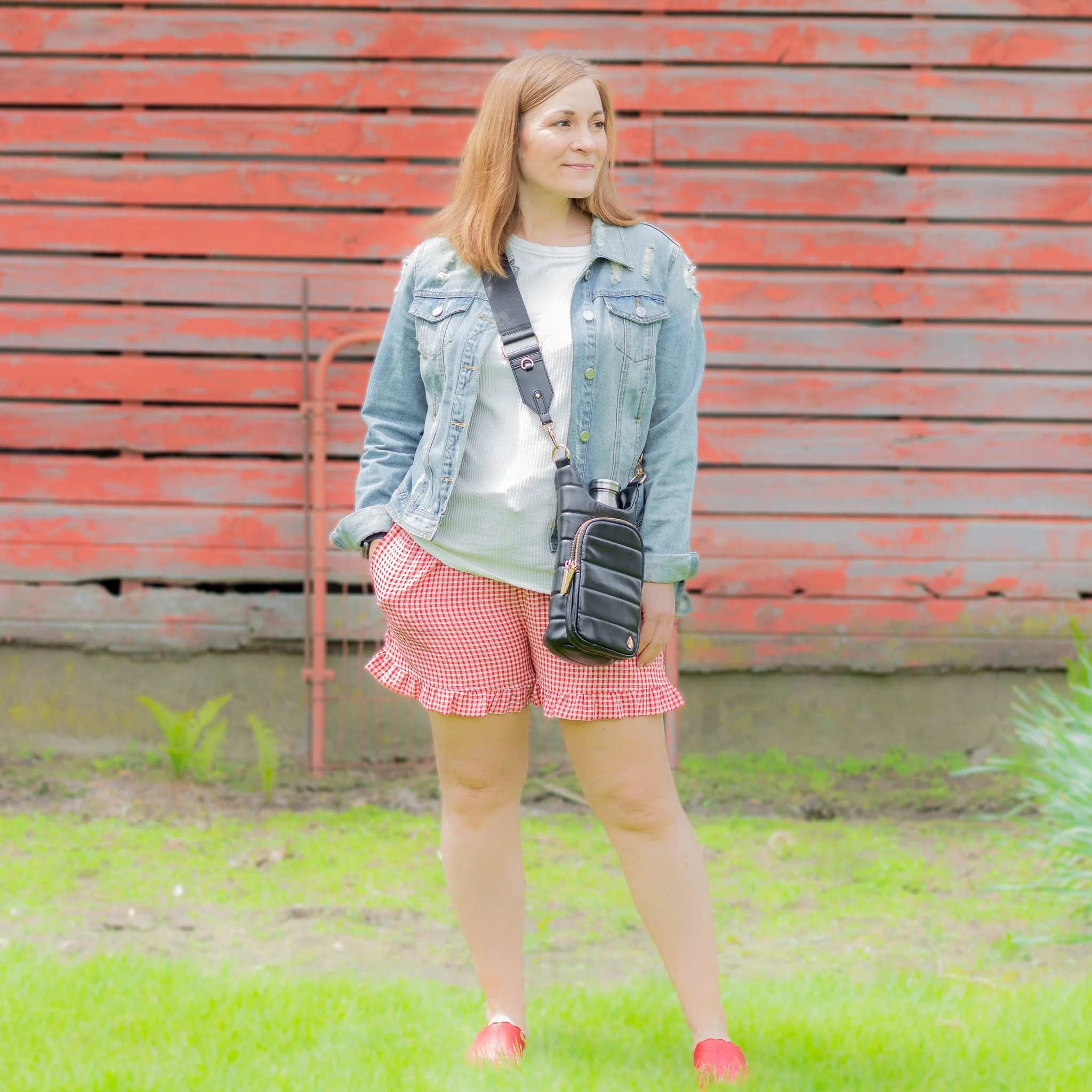 woman standing in front of barn with hand in pocket wearing red gingham shorts, white tee, denim jacket and black crossbody bag.