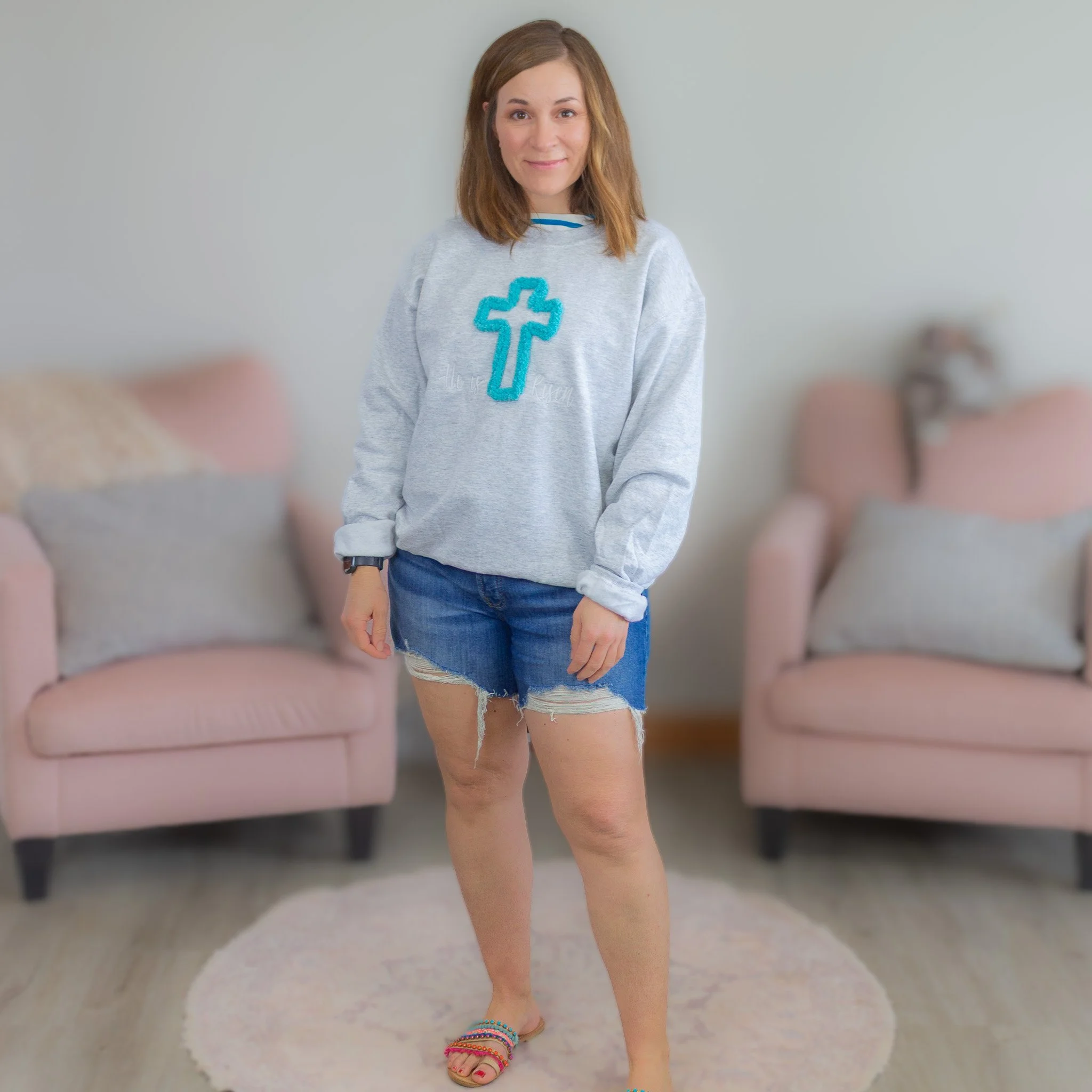 woman smiling wearing gray sweatshirt with teal chenille cross with words, "He is risen."