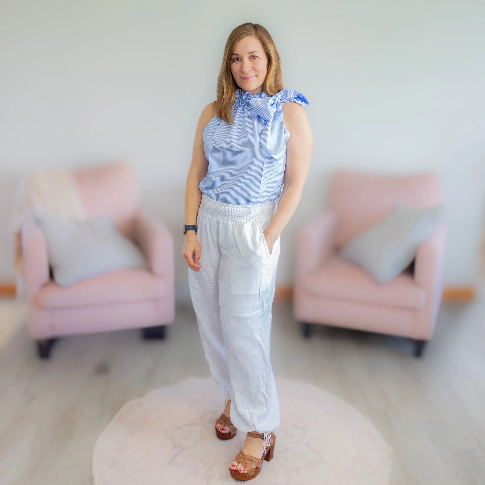 woman looking at camera wearing blue and white striped top, white pants and brown heeled sandals.